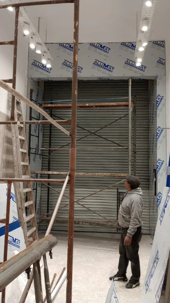 Construction site with a man in a hoodie and cap looking up at a closed metal rolling shutter, surrounded by scaffolding and insulation materials.