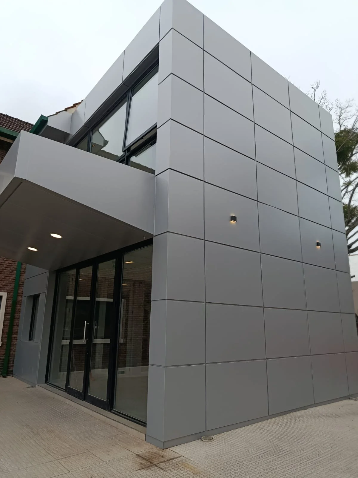 Modern building exterior with gray tiled facade, glass doors, and small wall-mounted lights.