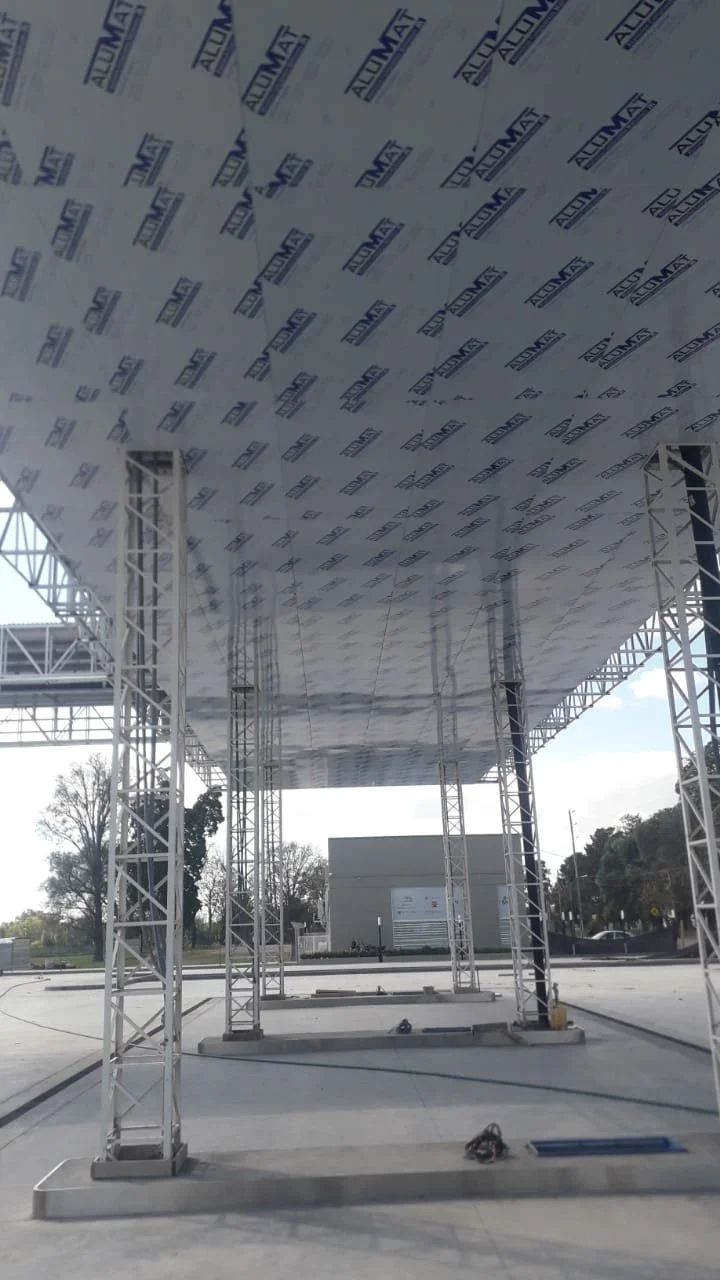 Under construction or installation site with metal scaffolding supporting a large, rectangular overhead structure. The structure is covered with a sheet printed with the logos 'Aluma' and 'Aluminum.' The background shows an outdoor area with trees and a building.