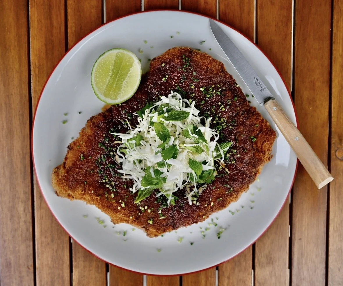 Breaded and fried fish fillet topped with shredded cabbage and herbs, served with lime wedge on a white plate with a knife on a wooden table.