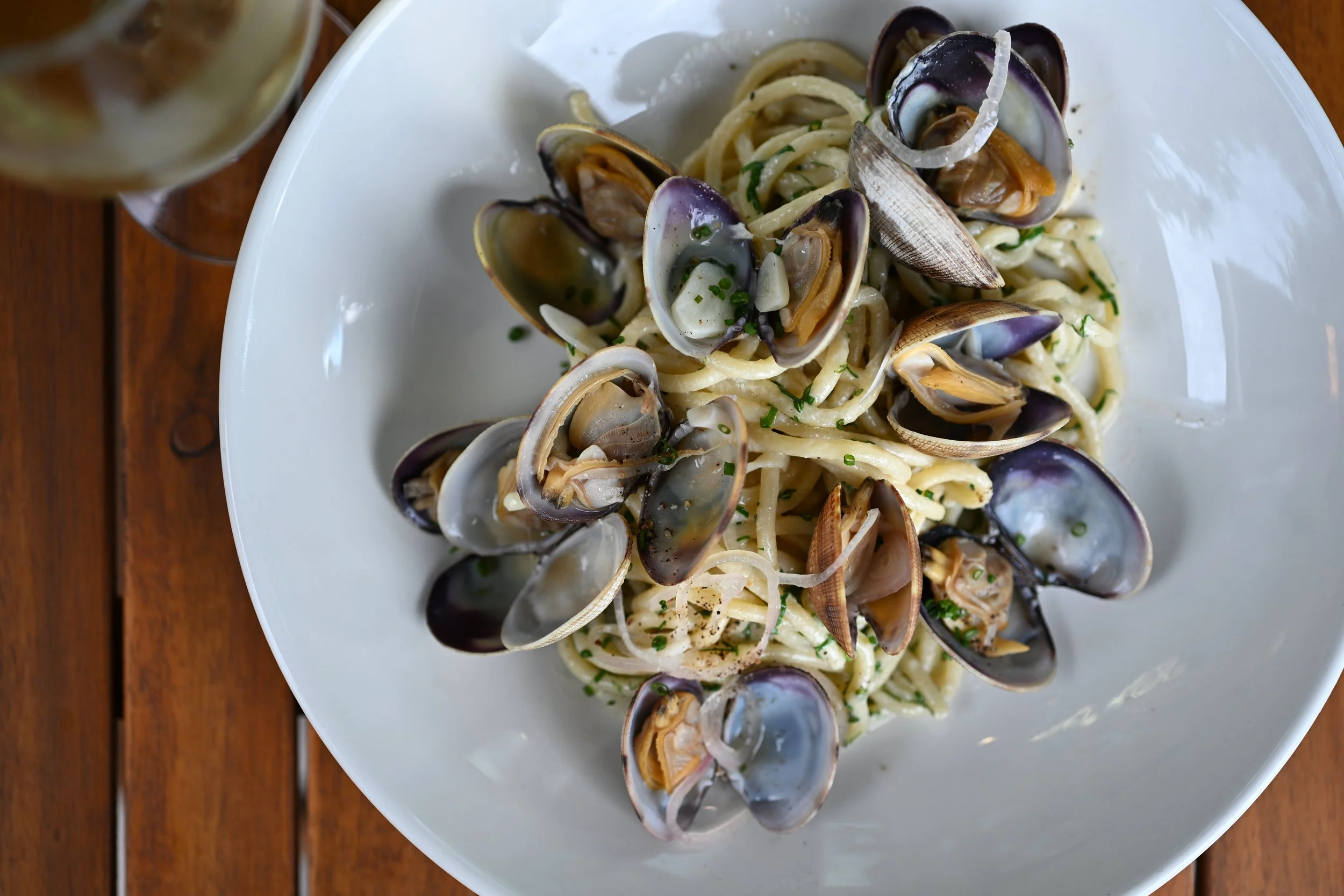 A white plate of spaghetti with clam sauce, garnished with chopped herbs, on a wooden table with a glass of white wine.