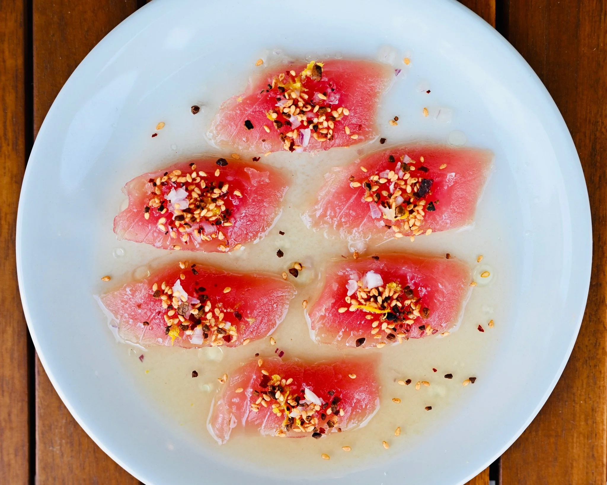 Plate with slices of raw tuna topped with chopped onions, chili flakes, sesame seeds, and lemon zest on a wooden table.