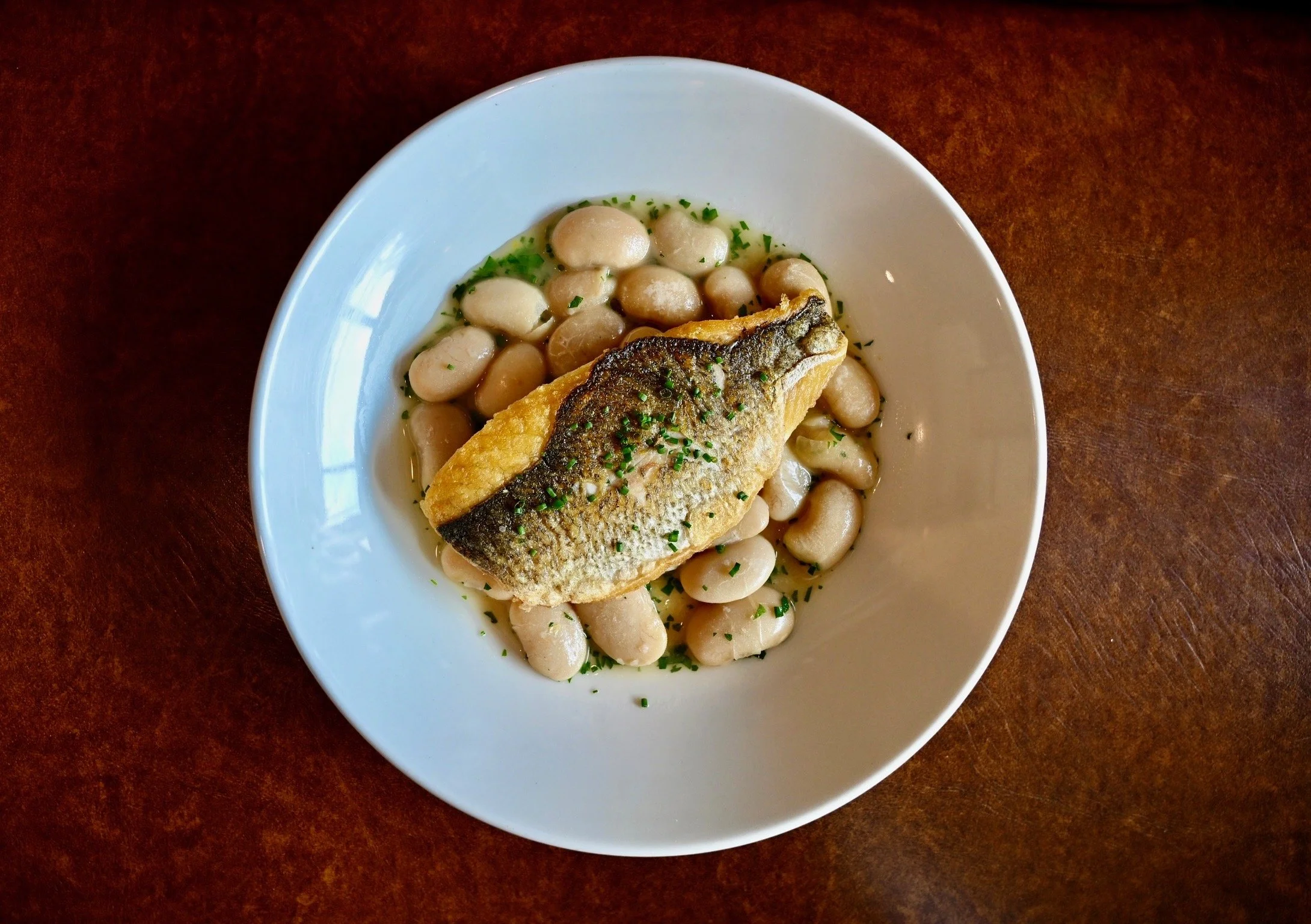 A white bowl containing a serving of cooked white beans topped with a cooked fish fillet garnished with chopped green herbs, set on a dark wooden surface.