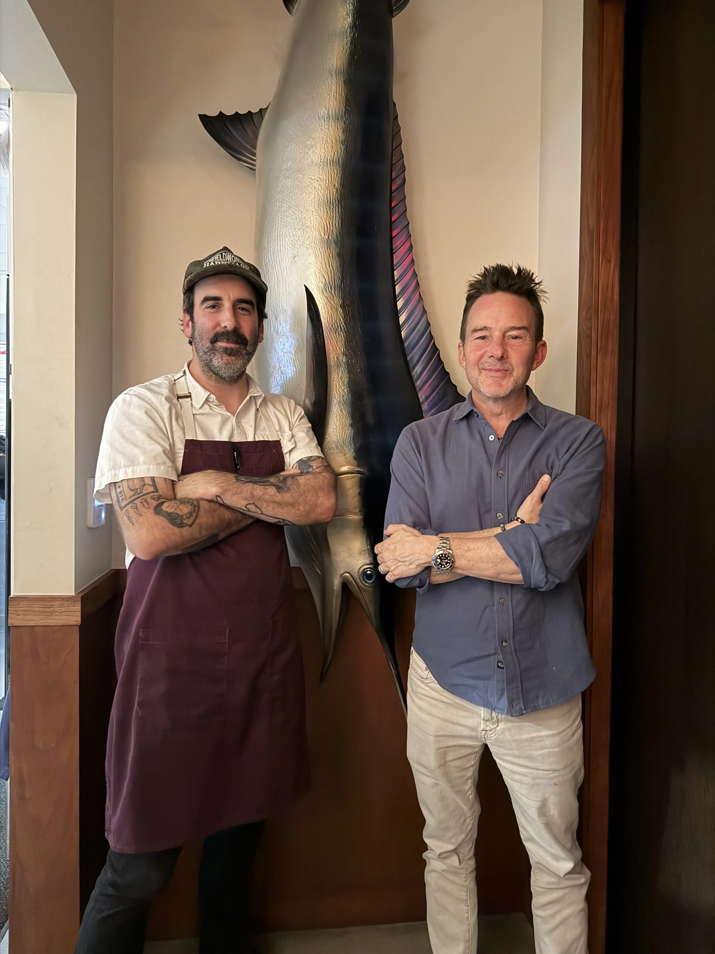 Two men standing next to a large fish mounted on the wall in a restaurant. The man on the left has dark hair, a beard, and is wearing a white shirt with a dark apron, and a hat. The man on the right has short dark hair, light skin, and is wearing a g