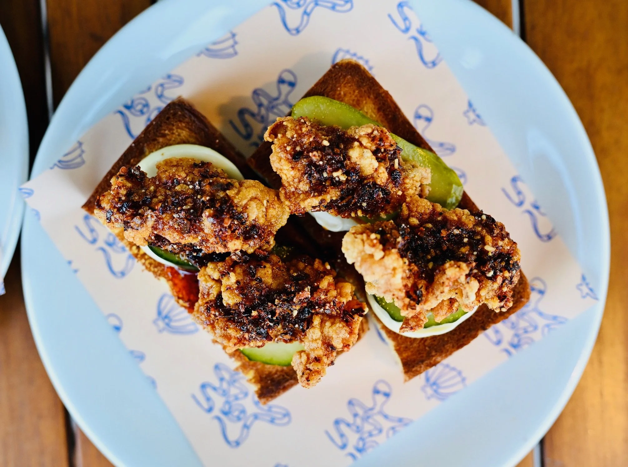 Fried chicken pieces topped with barbecue sauce on a sandwich with pickles, served on a bread slice with mayonnaise, on a white plate with blue writing.