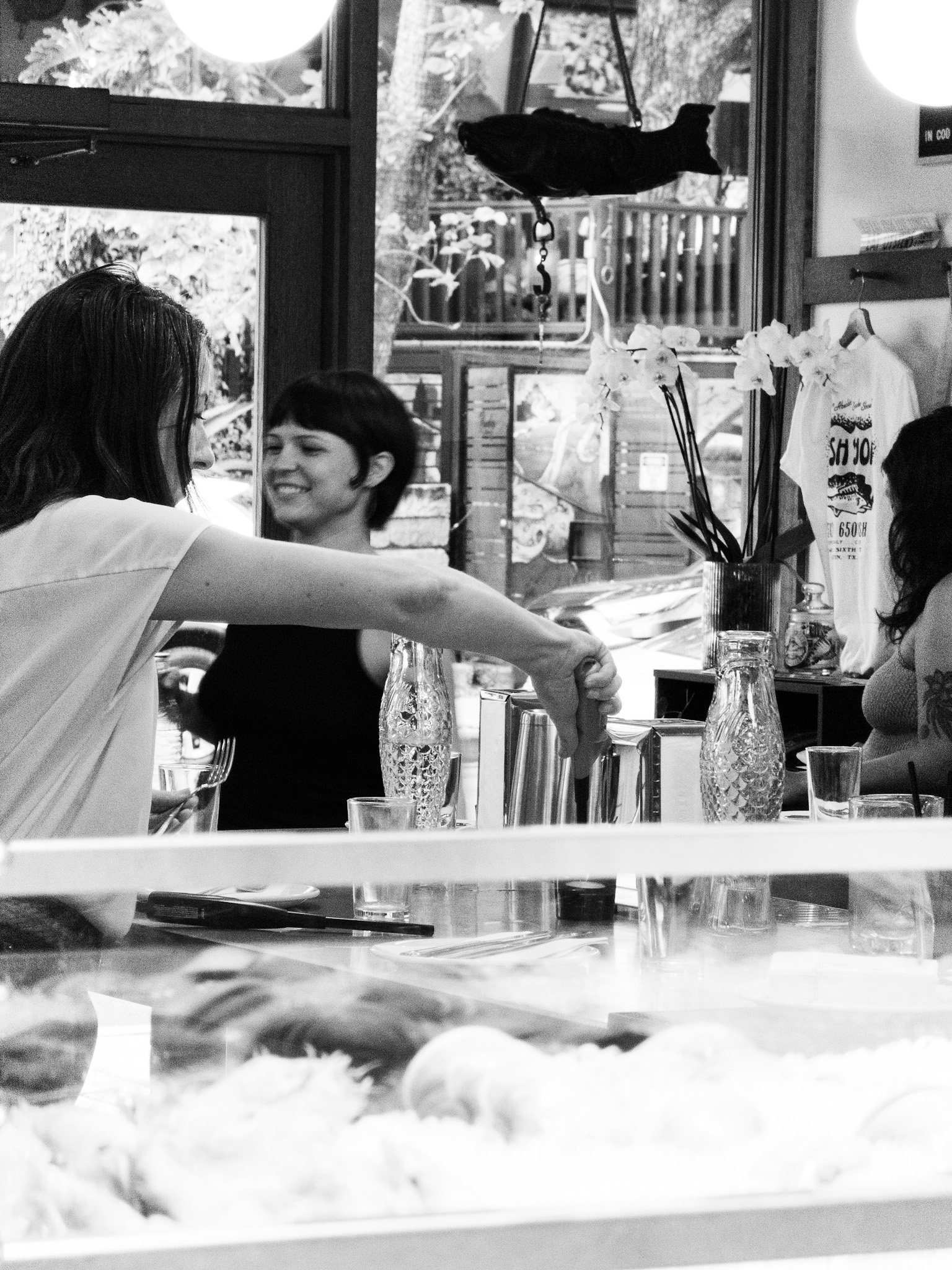 A black dog hanging upside down inside a store or café, looking out a window. Two women are sitting at a table; one arm reaches across, holding a glass. Bottles, plants, and items for sale are on the table, with a view of trees outside the window.
