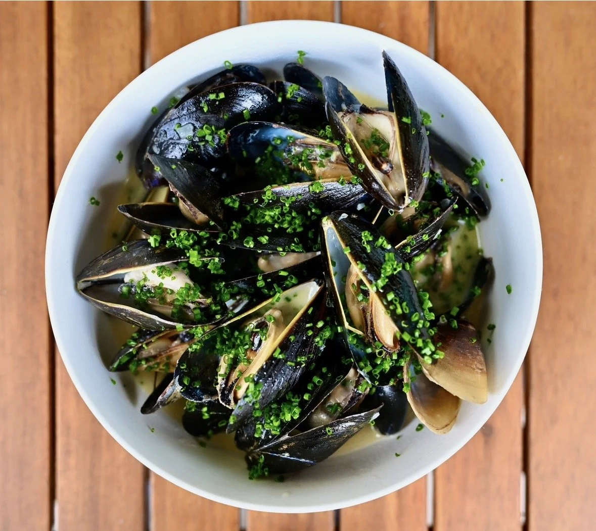 Bowl of cooked mussels garnished with chopped green herbs, on a wooden surface.
