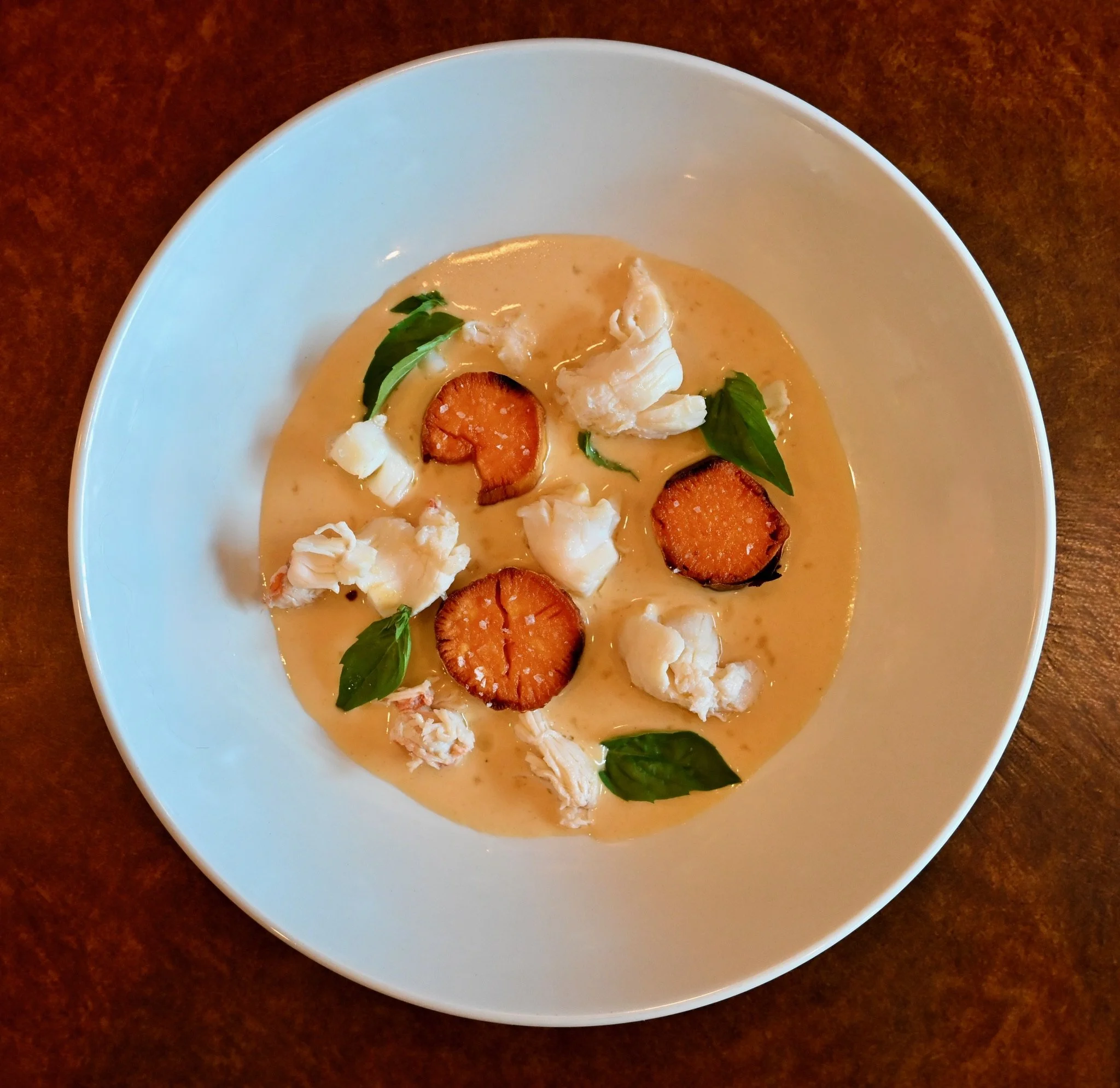 A white bowl with creamy soup, garnished with sliced carrots, greens, and cheese on a wooden surface.