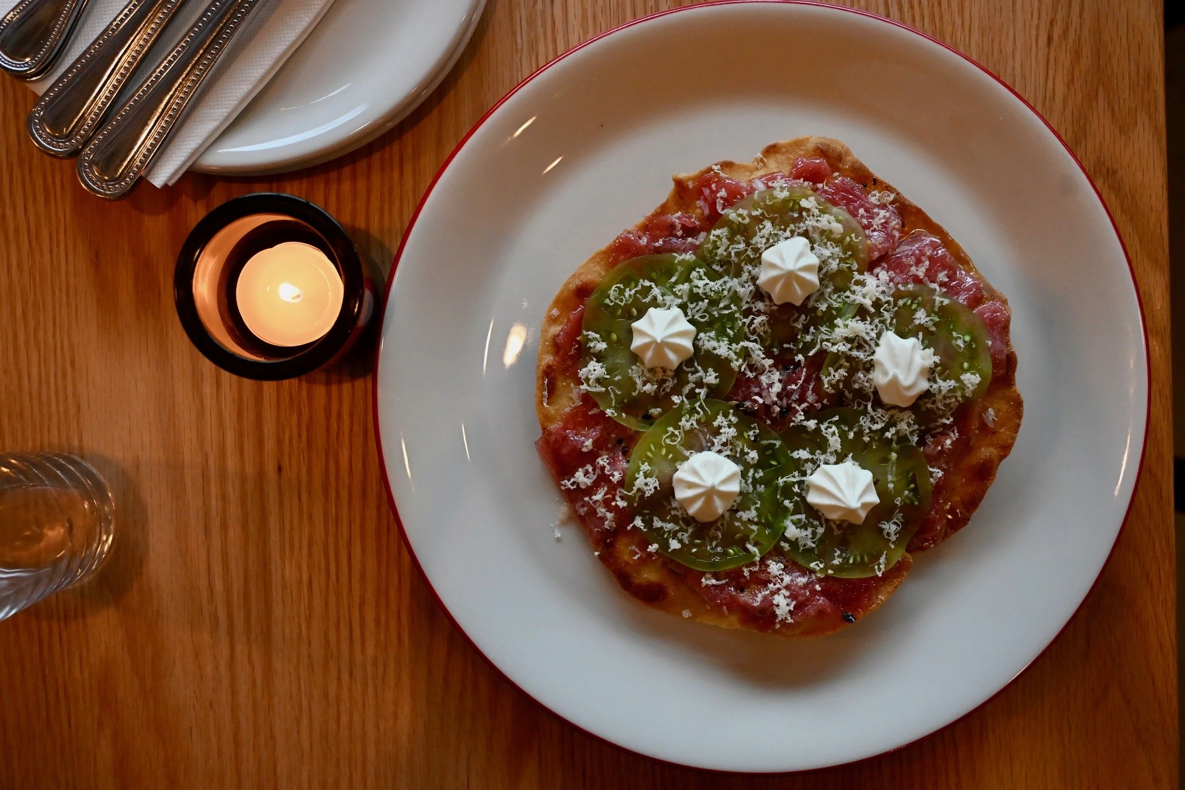A pizza topped with green bell peppers, shredded cheese, and dollops of sour cream on a white plate with a red rim, set on a wooden table with a small candle and silver utensils.