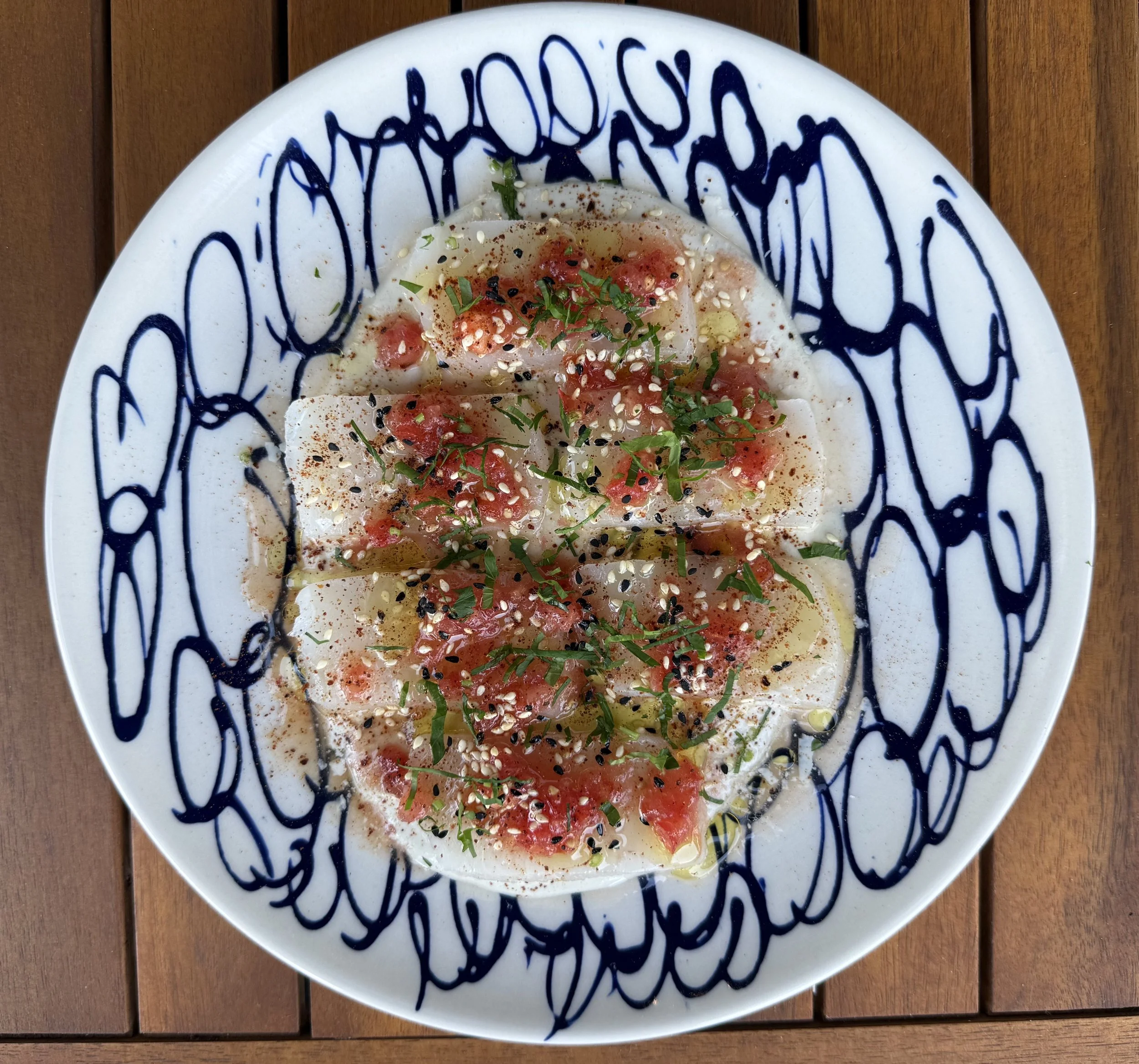 A white plate with blue abstract lines containing fish slices, garnished with green herbs, sesame seeds, and red seasoning, on a wooden table.