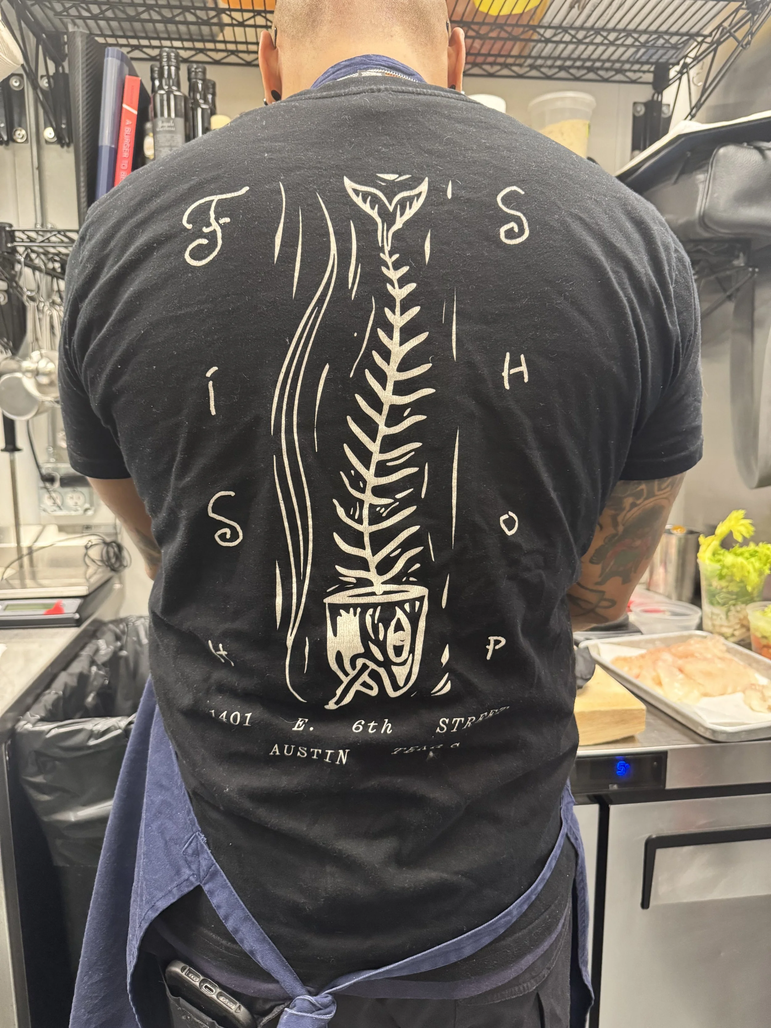 Back of a person wearing a T-shirt with a large snake illustration and text, in a kitchen or food prep area, with kitchen shelves and food containers in the background.