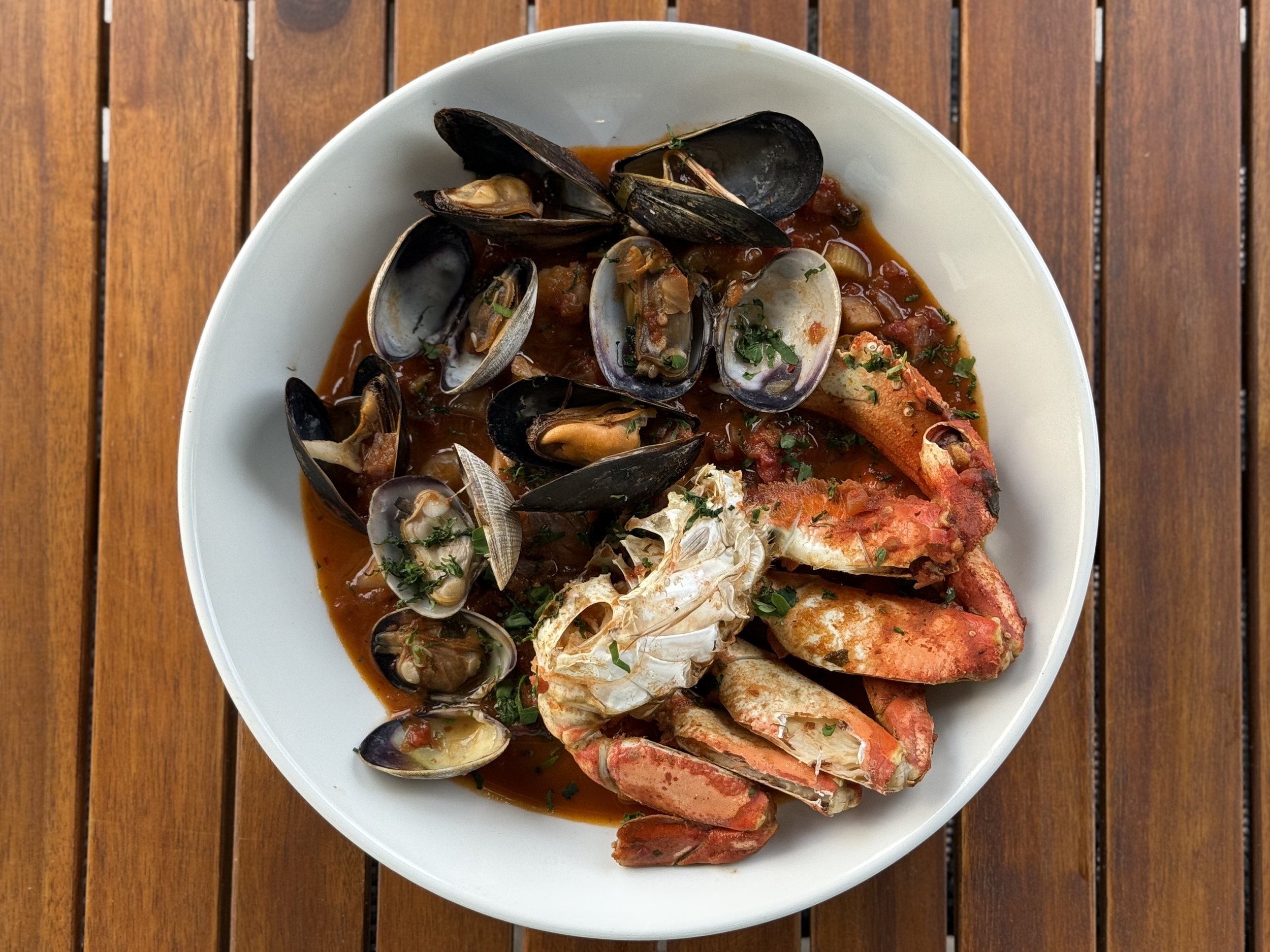 A white bowl containing a seafood stew with mussels, lobster, and other shellfish in a red broth garnished with herbs on a wooden table.