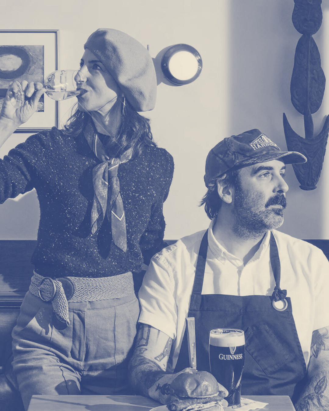 A woman in a beret and scarf drinks water while standing next to a seated man with a beard, wearing a cap and apron, in a restaurant with Guinness beer and a burger on the table.