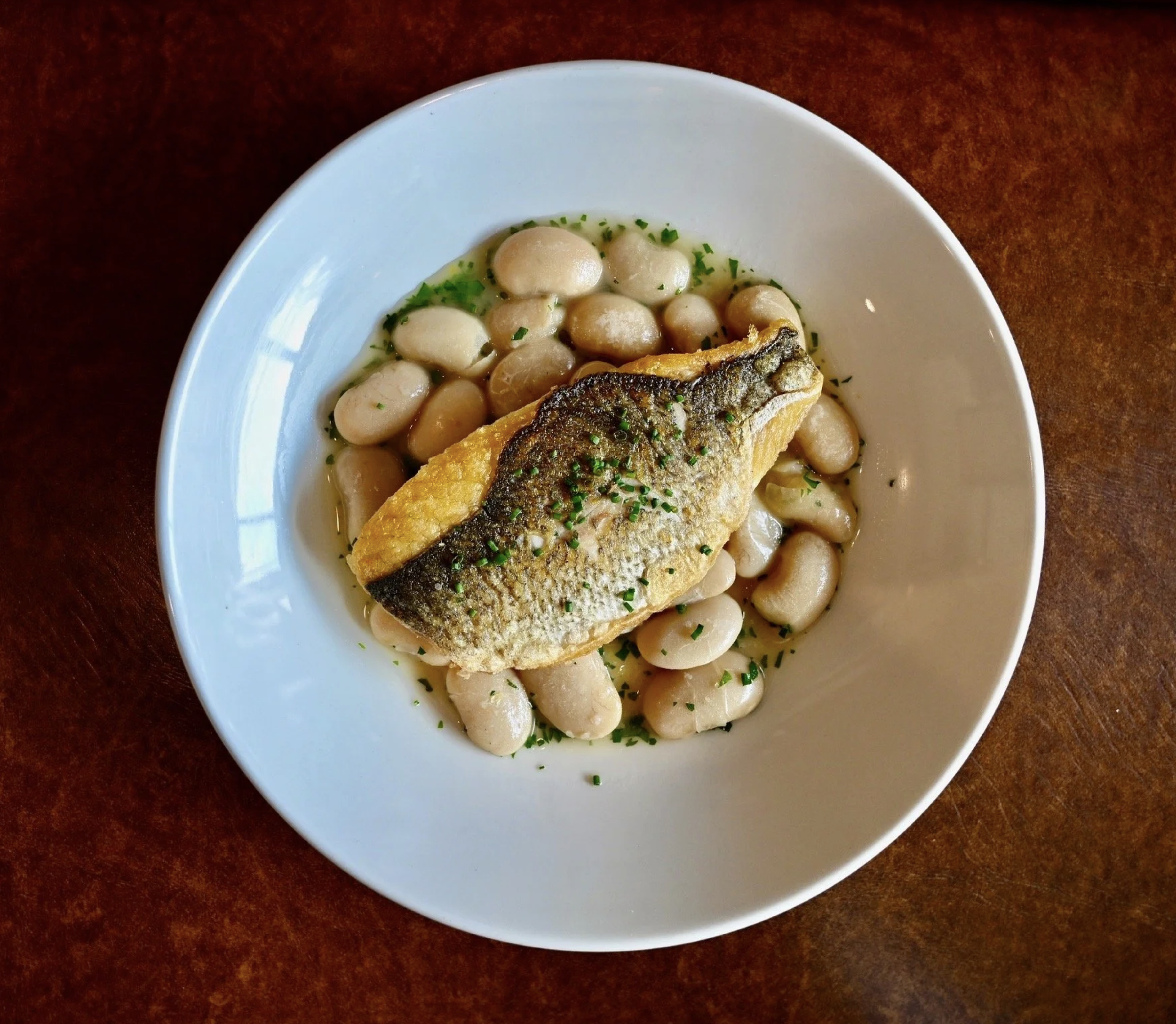 A white bowl containing cooked white beans garnished with chopped green herbs and a piece of fried fish with crispy, browned skin on top of the beans, on a dark wooden surface.