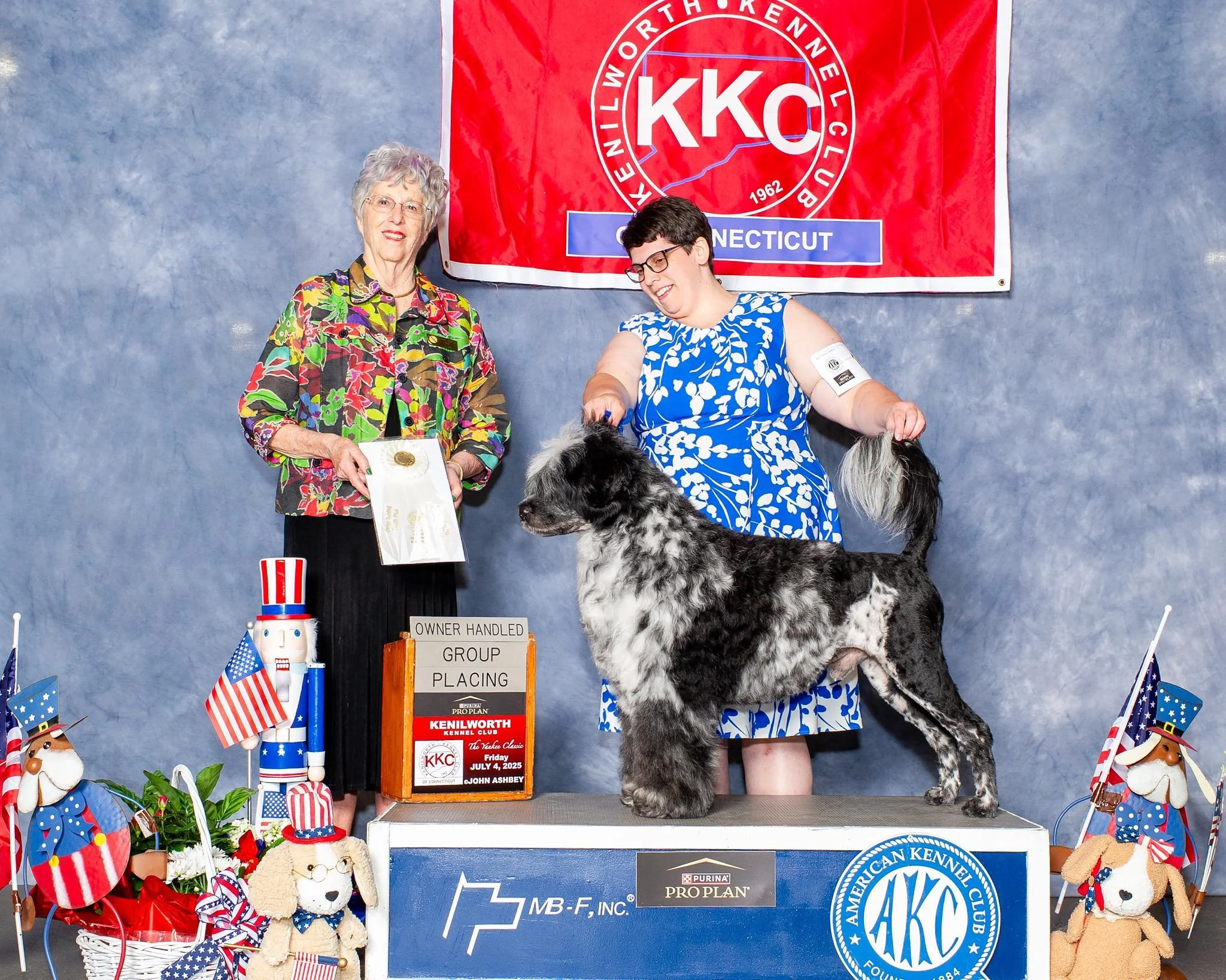 A woman with gray hair and a colorful floral jacket standing beside a woman with short dark hair, glasses, and a blue and white patterned dress holding a large black and white dog at a dog show. They stand in front of a red and white banner with dog club logos, and the dog is standing on a platform. Decorative items including flags, plush dogs, and ribbons are displayed around them.