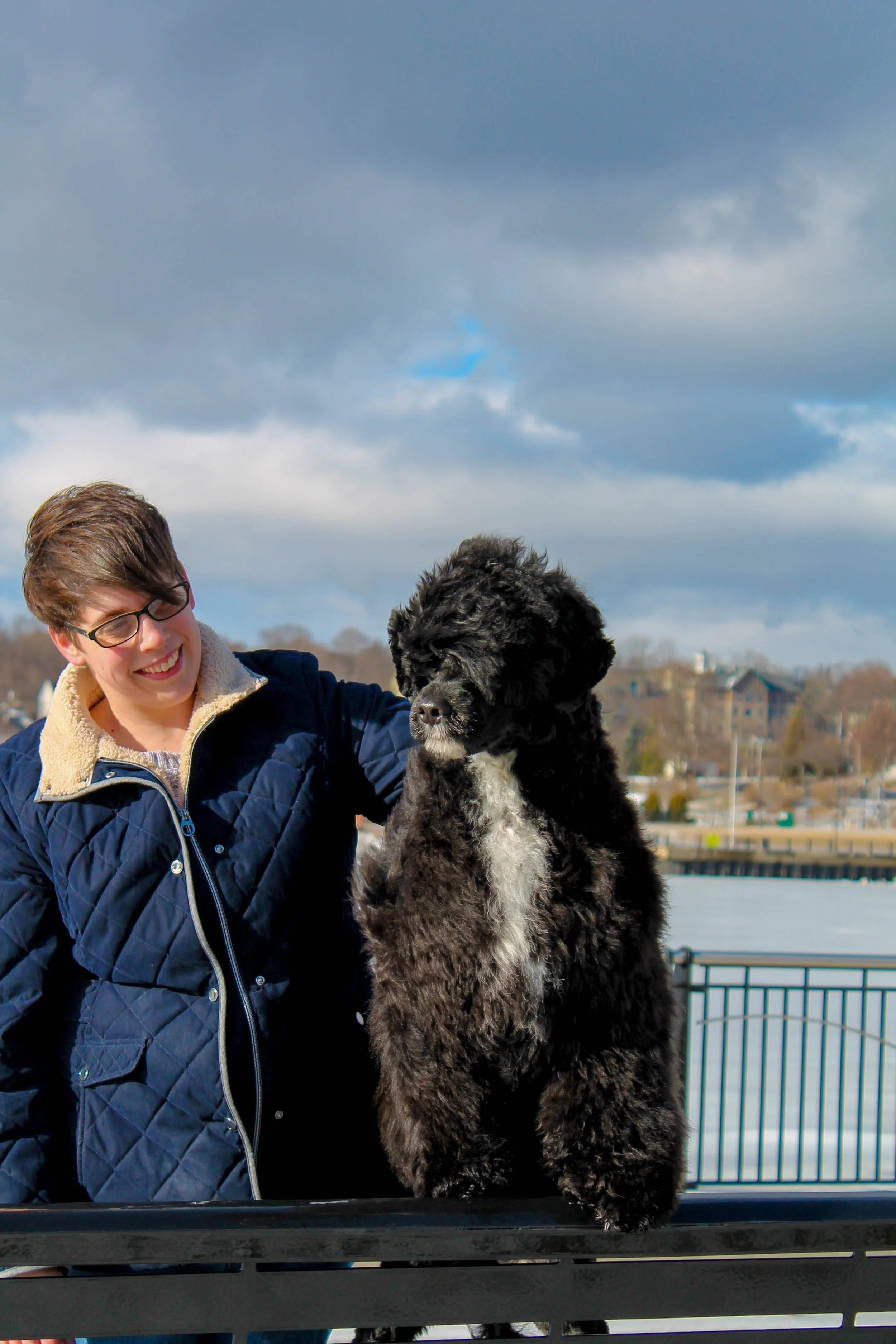 A woman with short brown hair, glasses, and a blue jacket is smiling and holding a large, curly black and white dog outdoors near a body of water and a cityscape with cloudy skies in the background.