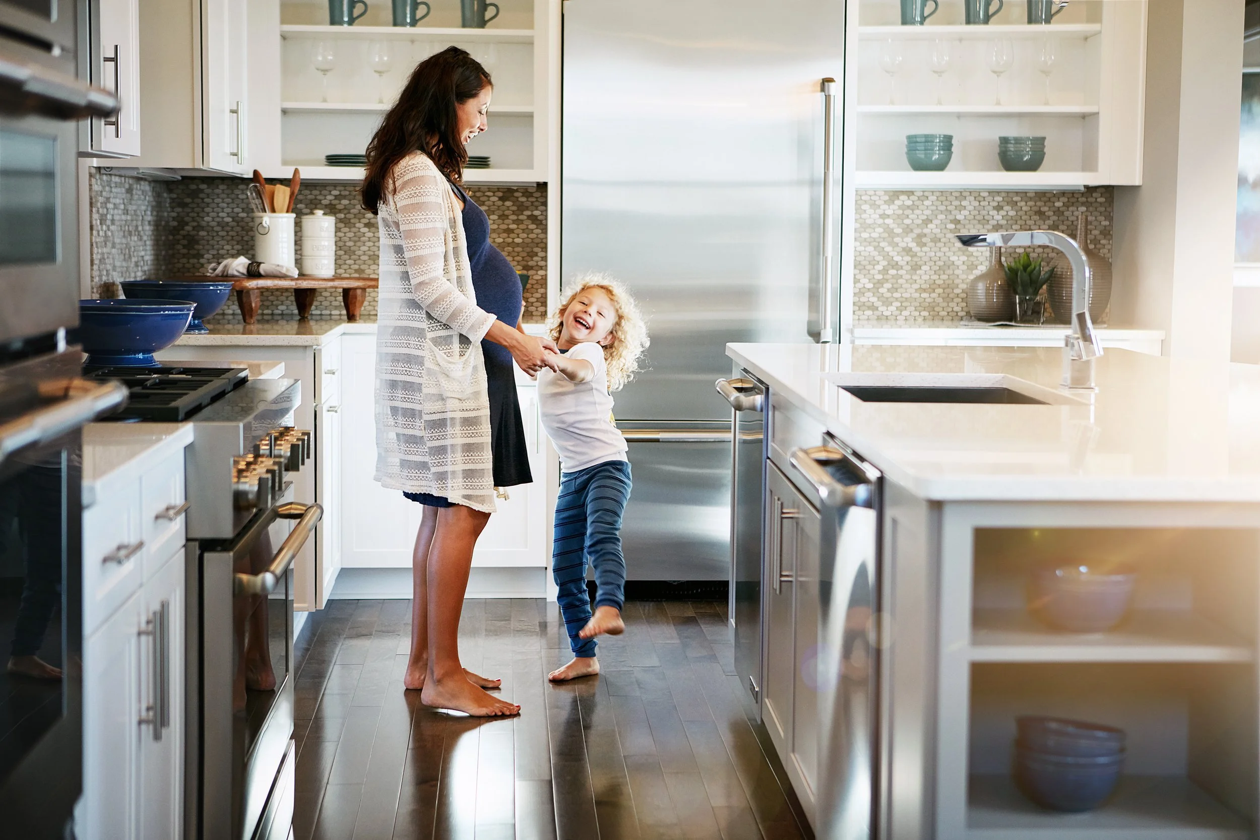 A woman and a young girl are happy and smiling in a modern kitchen, holding hands and playing together. The woman is in a maternity dress, and the girl is barefoot, wearing blue striped pants. The kitchen has white cabinets, stainless steel appliances, and dark wood flooring.