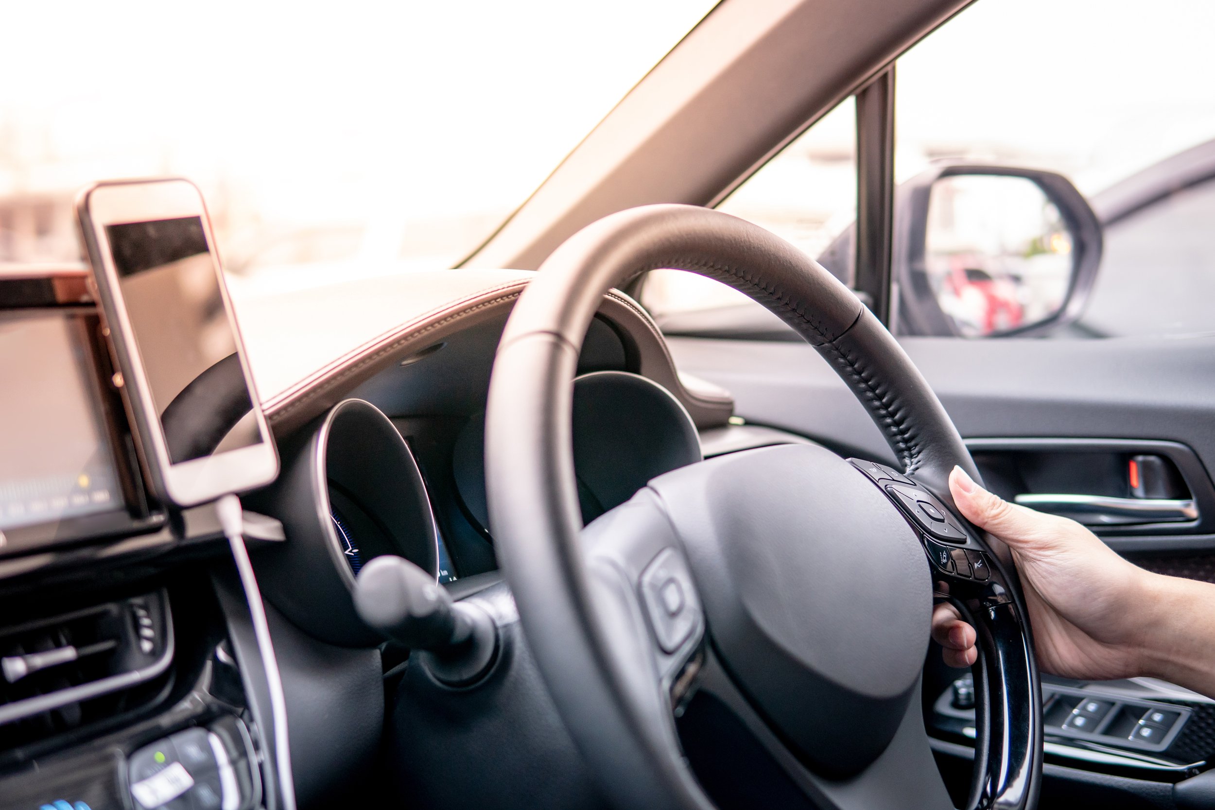 Inside view of a modern car dashboard, steering wheel, and a person's hand operating the controls, with a mounted smartphone on the dashboard.