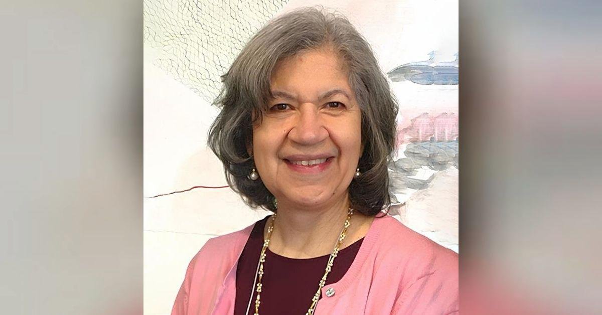 A smiling middle-aged woman with shoulder-length gray hair, wearing a pink shawl, a dark top, pearl earrings, and a gold necklace, standing indoors in front of a pastel painting.