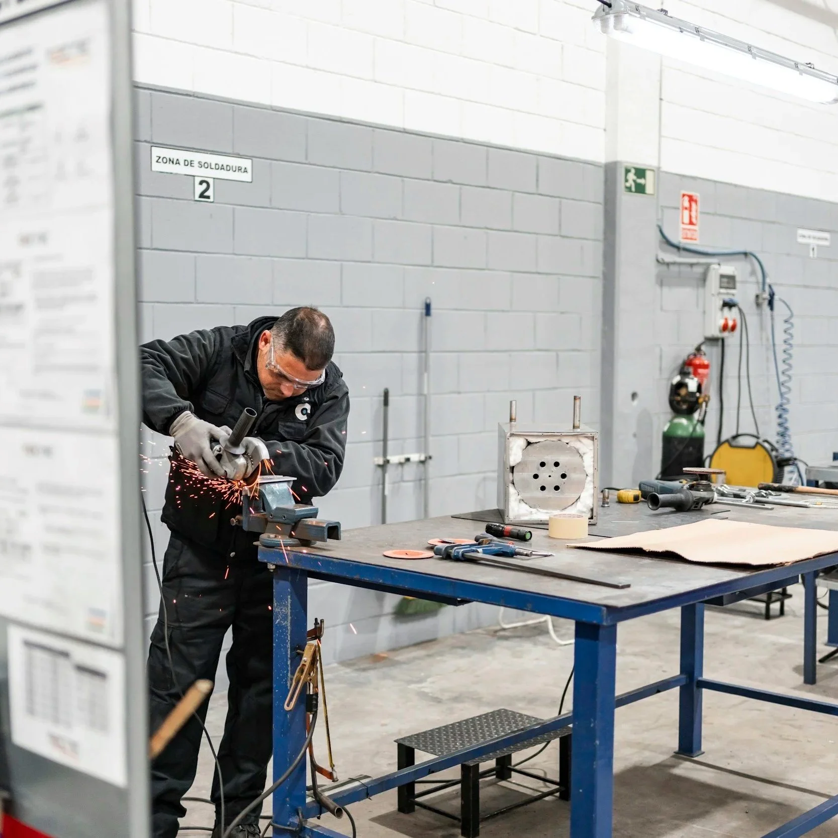 A man wearing safety glasses Welder in a workshop, using an angle grinder with sparks flying. The workshop has a gray cinder block wall and various tools and equipment on a blue workbench.