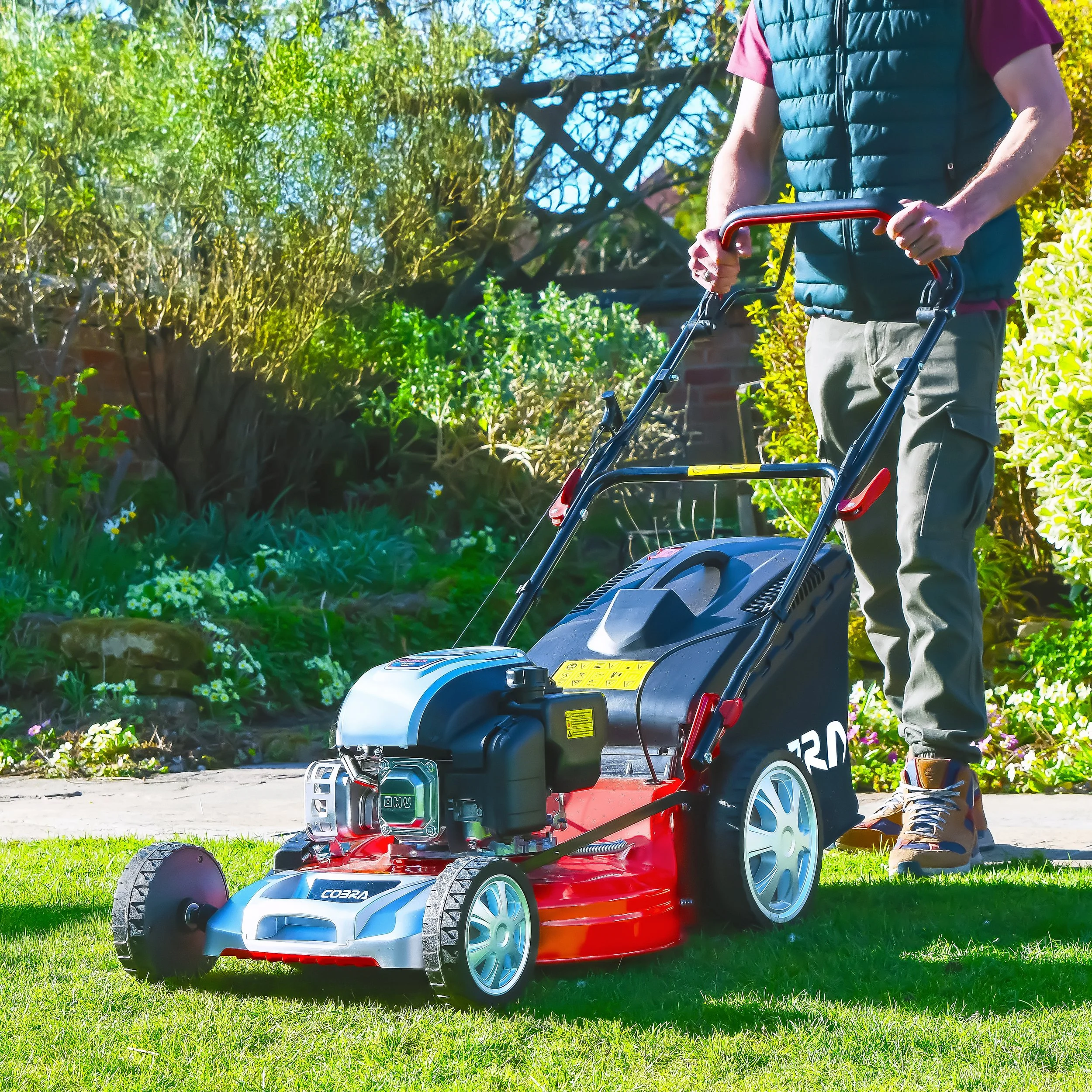 Man using a gas-powered lawn mower in a green garden with trees and bushes.