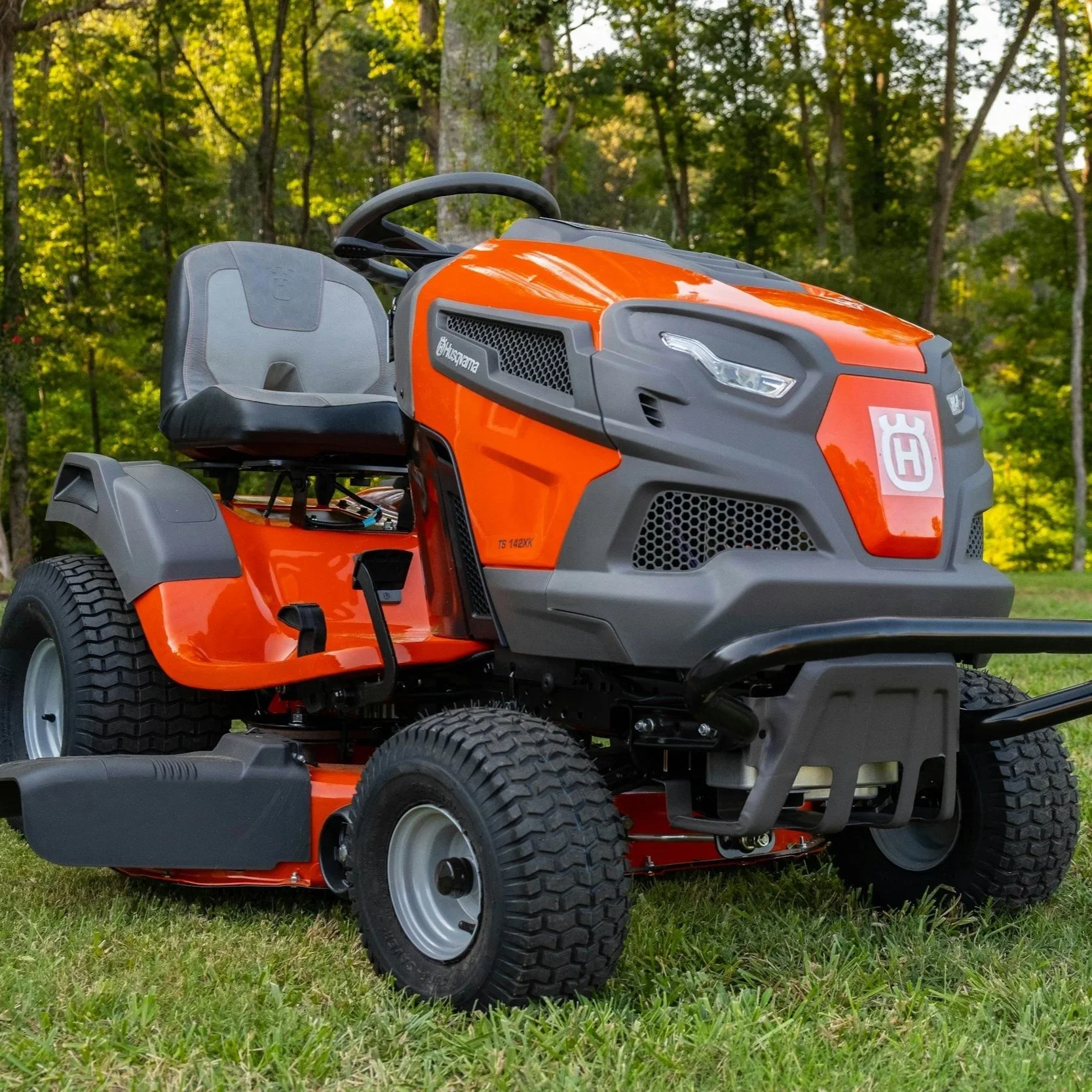 Close-up of an orange and gray Husqvarna riding lawn mower on a grassy area with trees in the background.