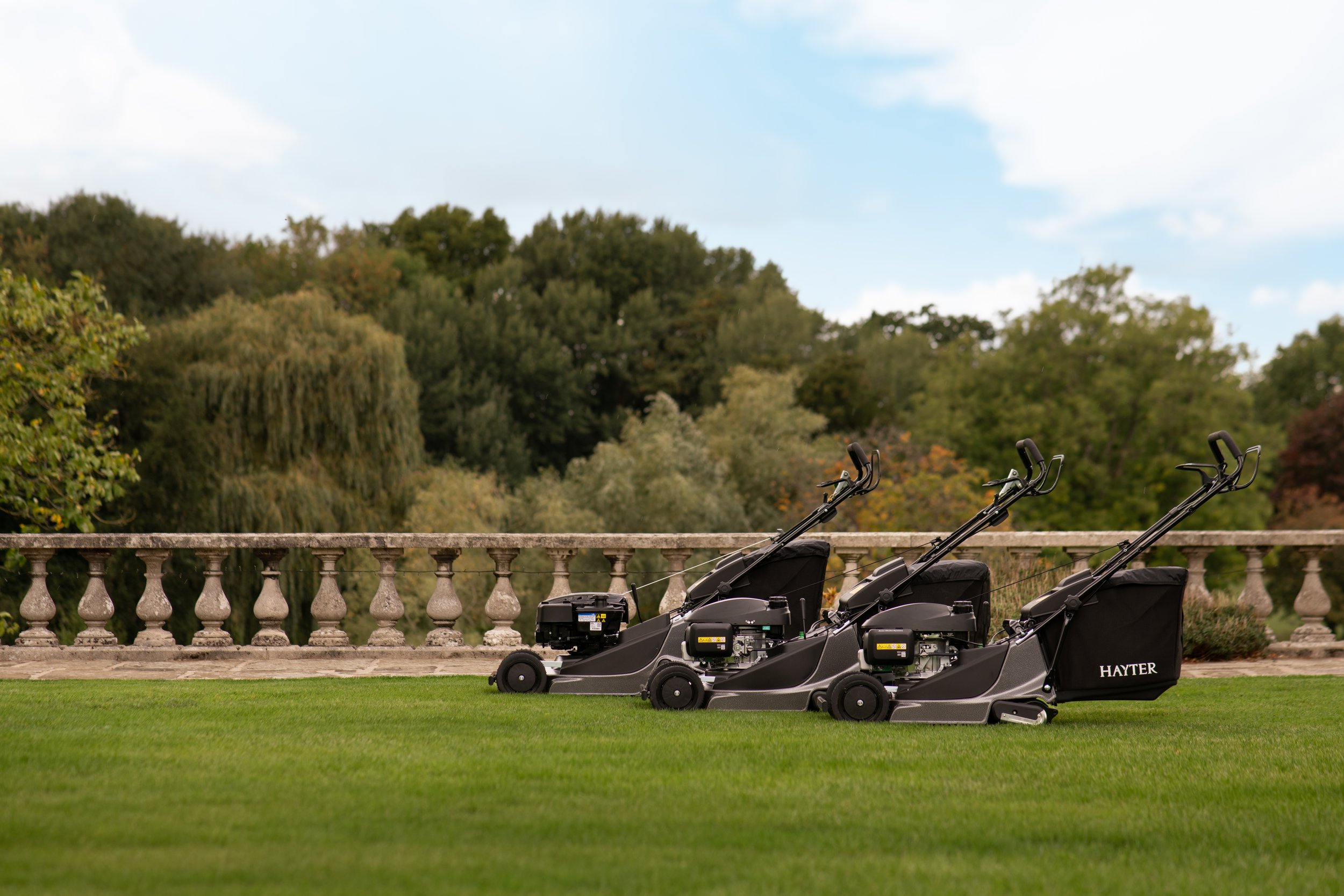 Four black lawn mowers lined up on a grassy area outdoors, with trees and a stone railing in the background.