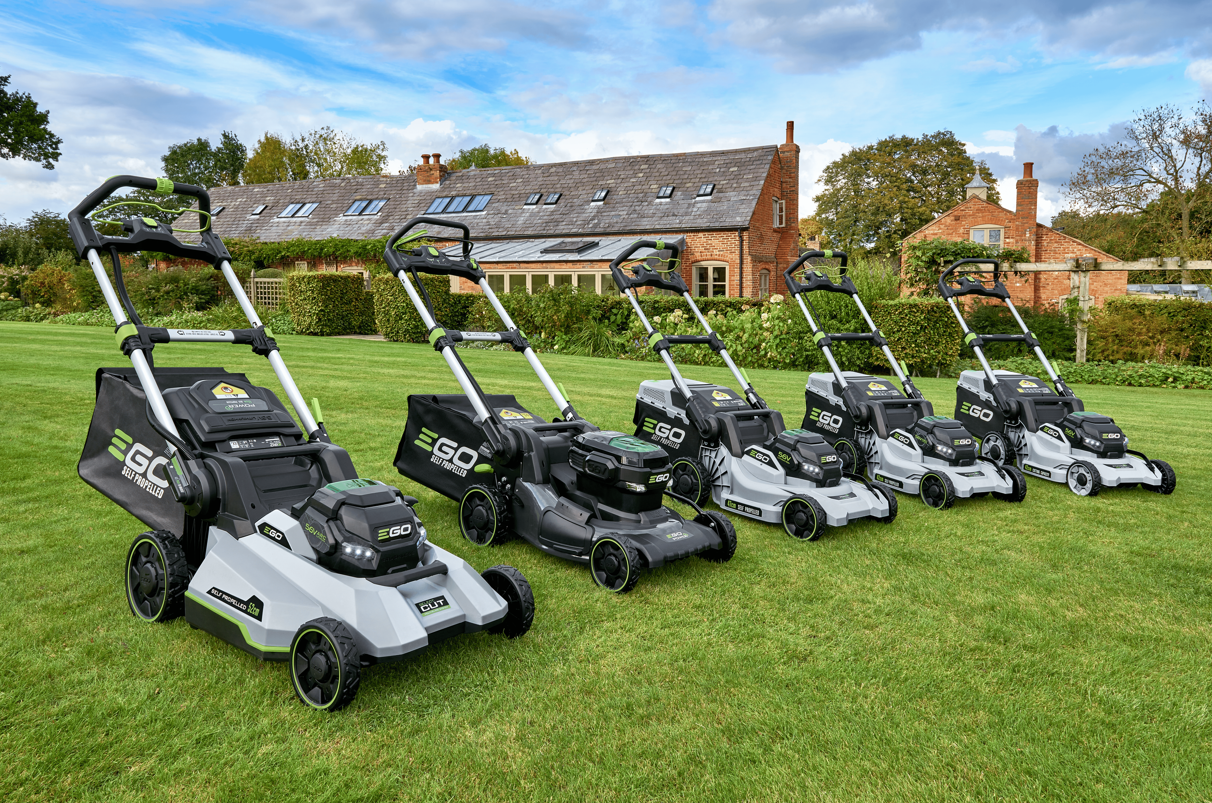 Line of self-propelled lawn mowers on a well-manicured lawn in front of a brick house with a garden.
