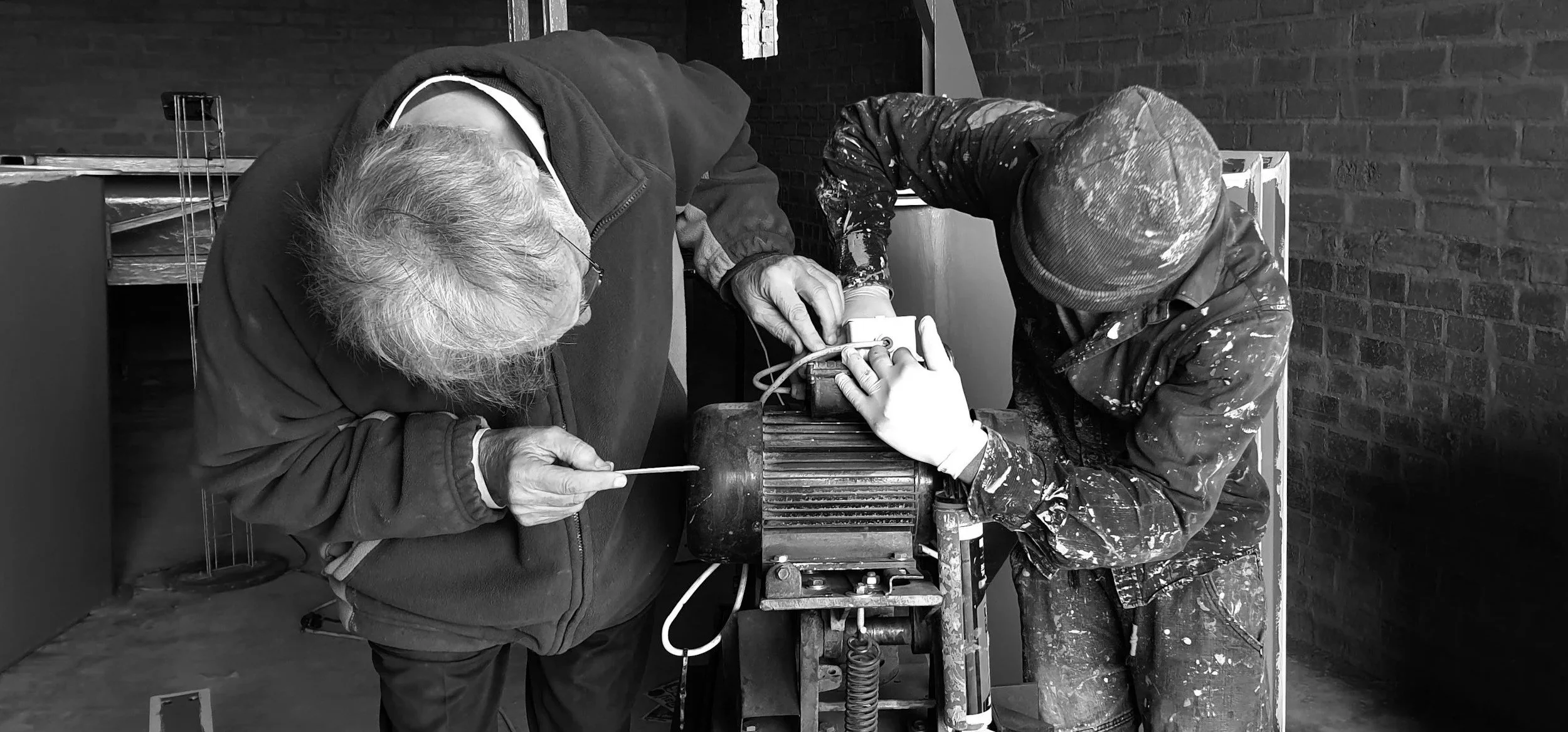 Two people working together on a mechanical project, one older with glasses and the other younger wearing a beanie, in a workshop with brick walls.