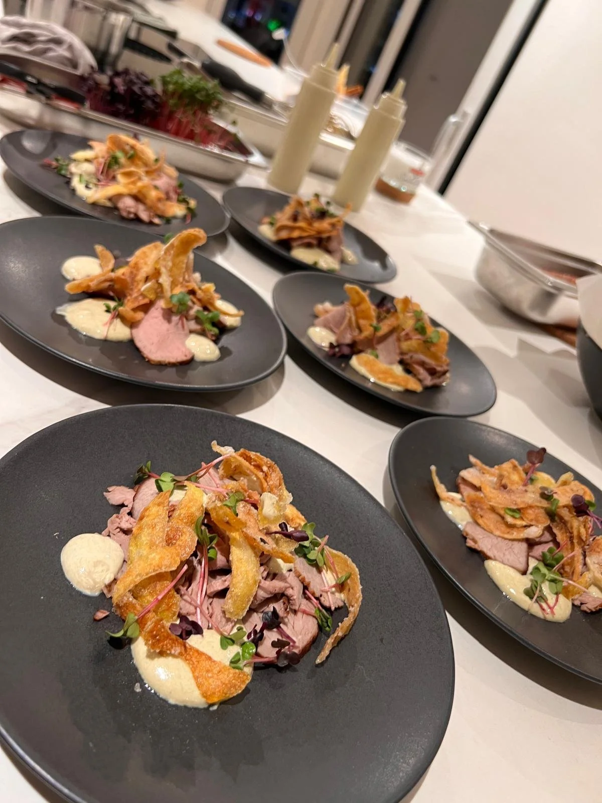 Several black plates with gourmet dishes featuring slices of cooked meat, crispy fried garnish, microgreens, and dollops of creamy sauce on a white table in a kitchen setting. Düsseldorf Köln Leverkusen Fußball Football Private Chef Private Meal Prep