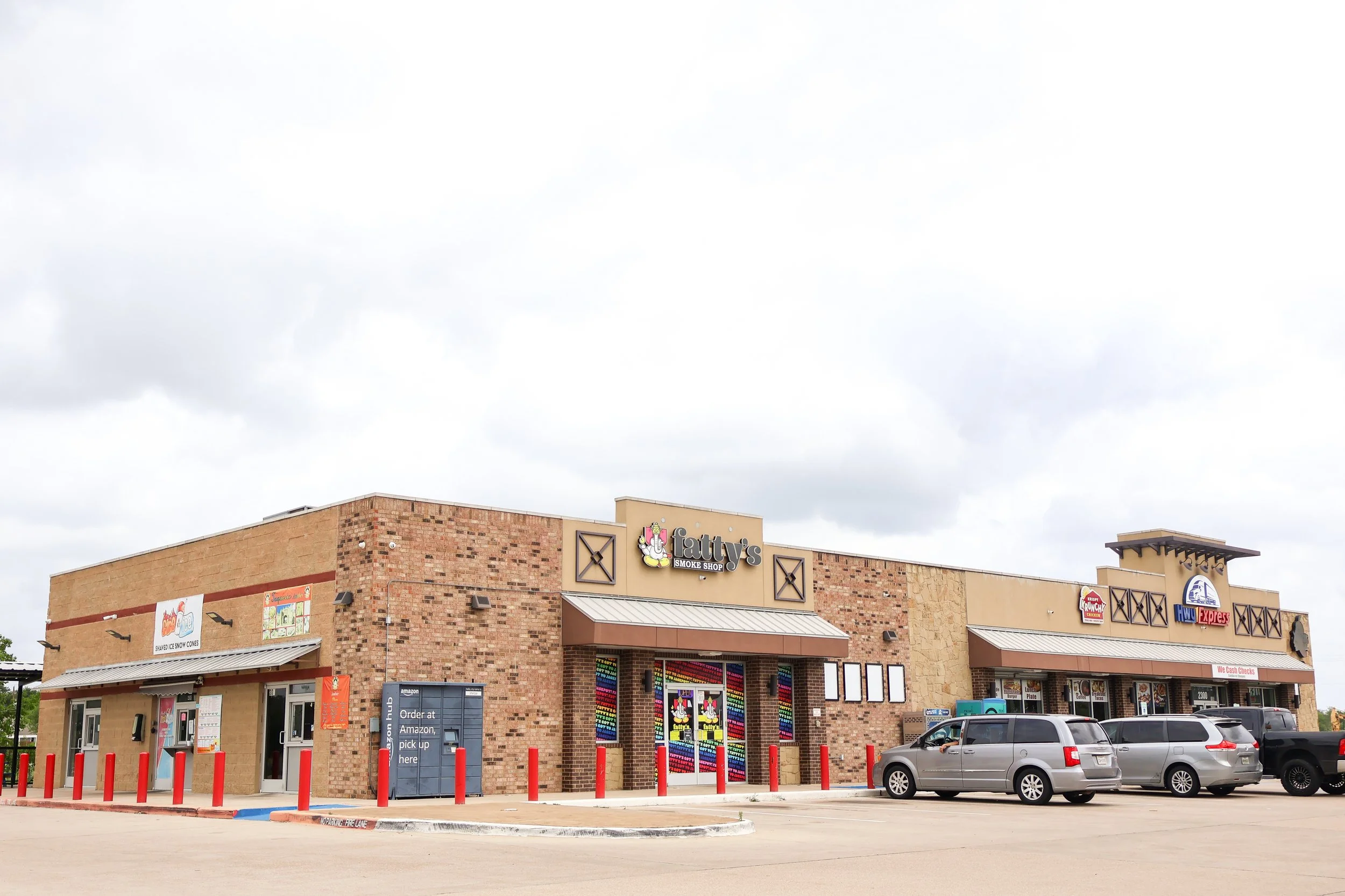 A shopping center in Bryan, Texas off Hwy 21 with a brick facade featuring various store signs, including Fatty's Smoke Shop. There is a parking lot with several cars and red parking barriers, and an overcast sky overhead.