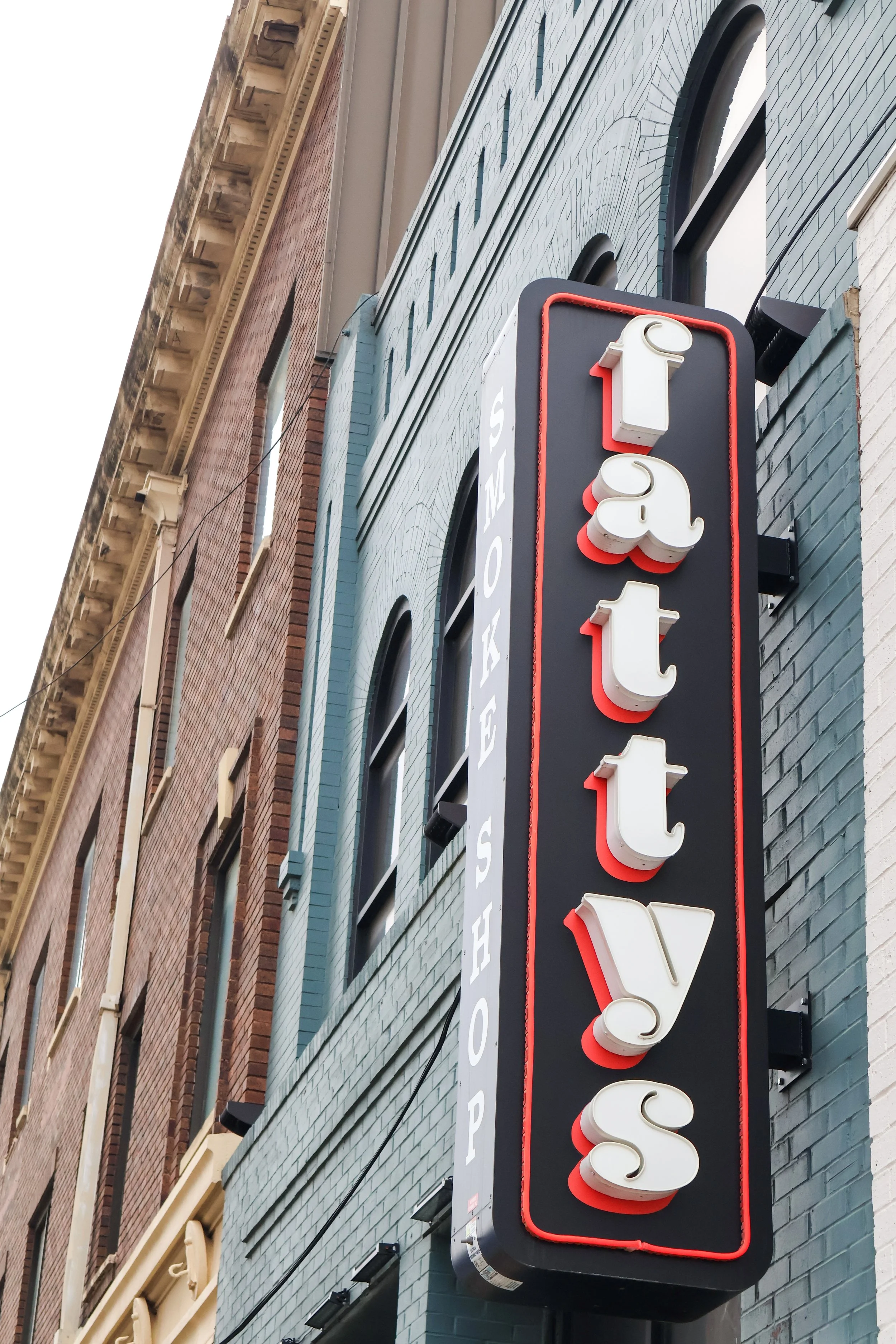 Vertical sign with the word "Fatty's" in 3D white letters outlined in red, mounted on a brick building with arched windows.