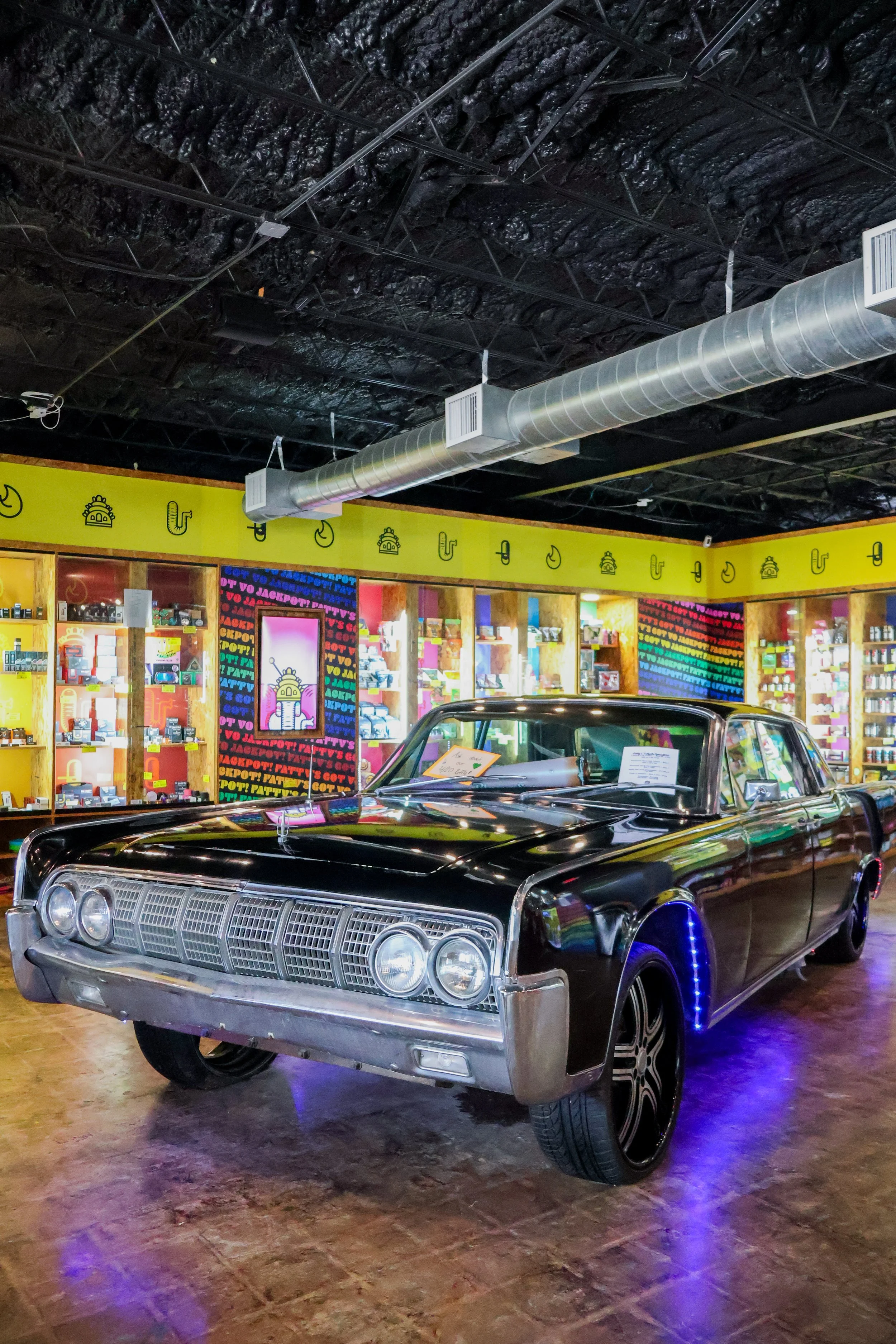 A black vintage car with chrome details is on display indoors at the Fatty's Smoke Shop location in Bryan, Texas, illuminated by purple and blue lights.