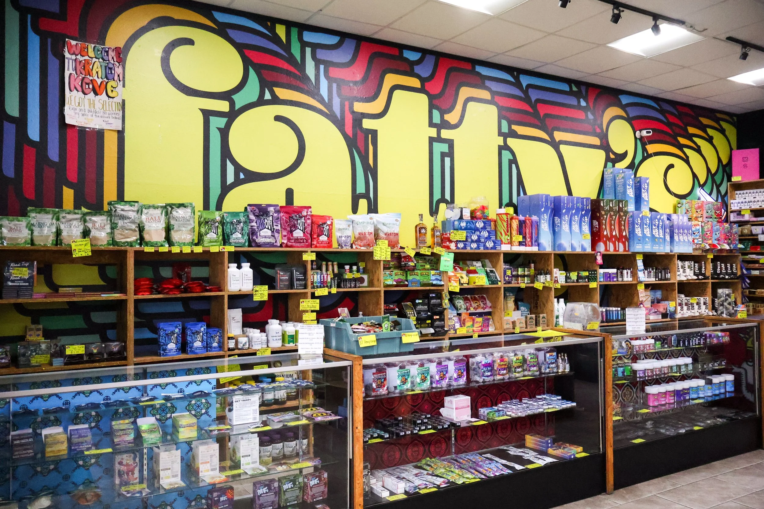 A store shelf displays various smoke related products. The wall behind features a large, colorful graffiti-style mural with yellow, red, blue, green, and black patterns.