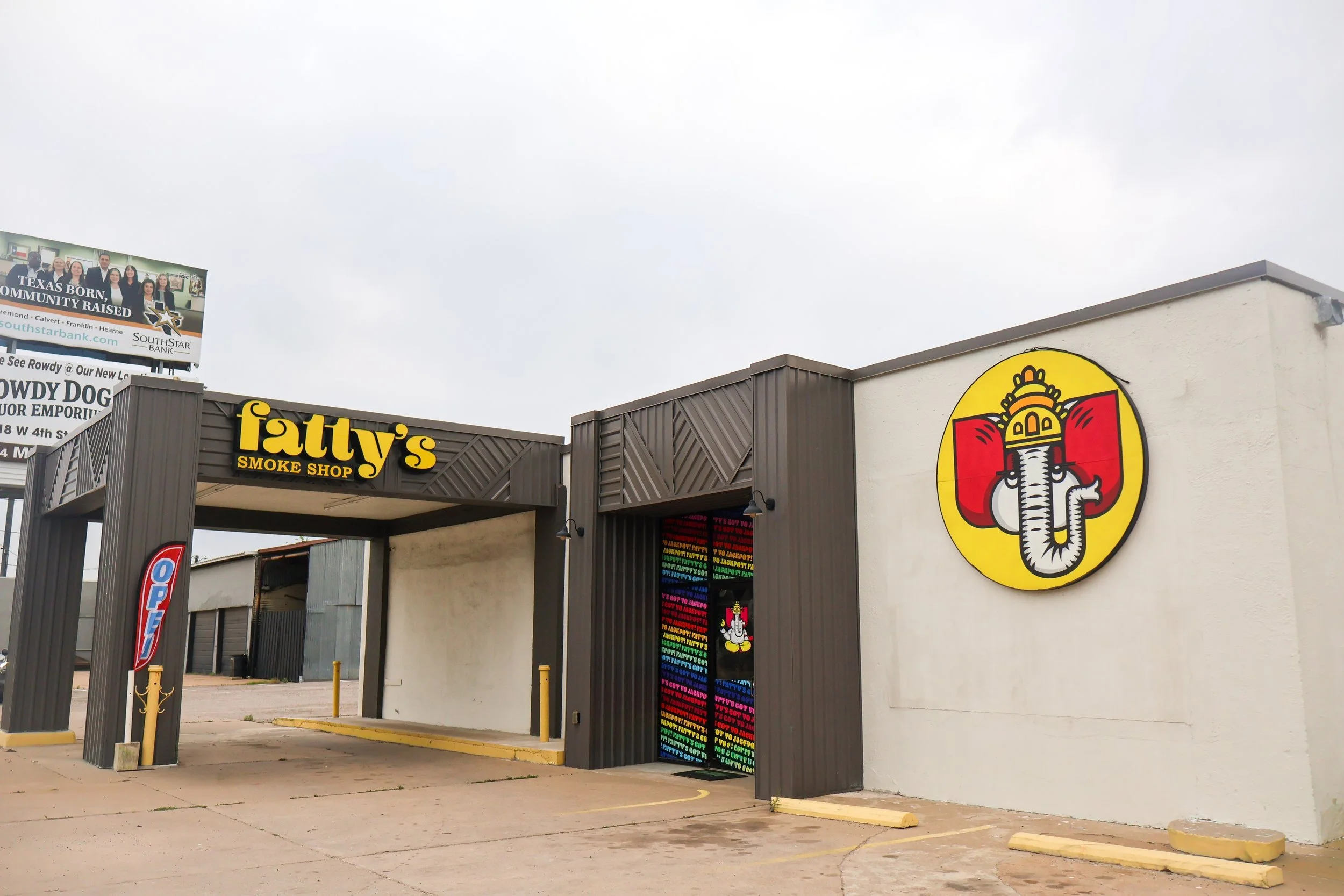 An entrance to a smoking shop called Fatty's Smoke Shop with a large colored logo of an elephant's head on the wall, a colorful vertical banner, and a billboard in the background.
