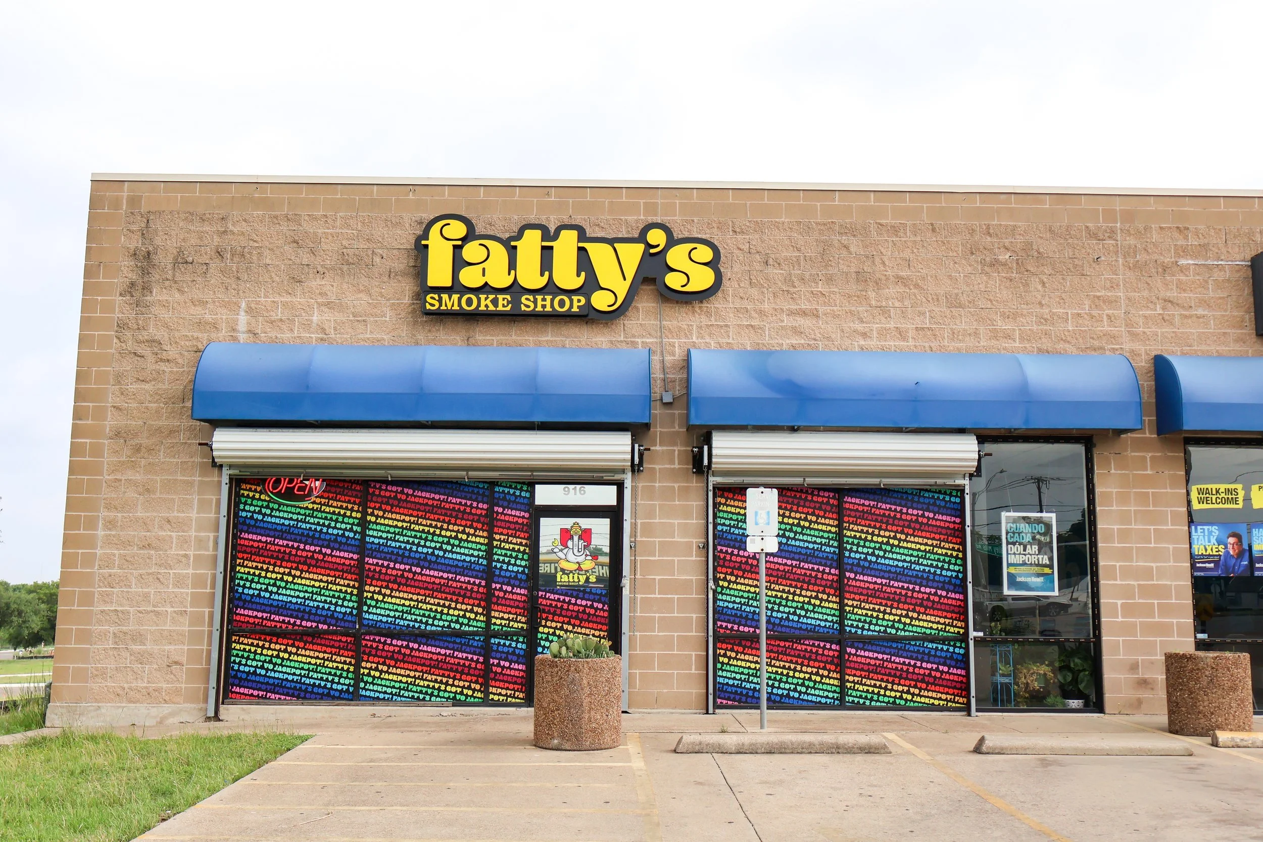 Exterior of a smoke shop in Waco, Texas, with a yellow sign reading 'Fatty's SMOKE SHOP'. The storefront has two large colorful posters, and a blue awning. There are small potted plants outside, and the pavement includes parking spaces.