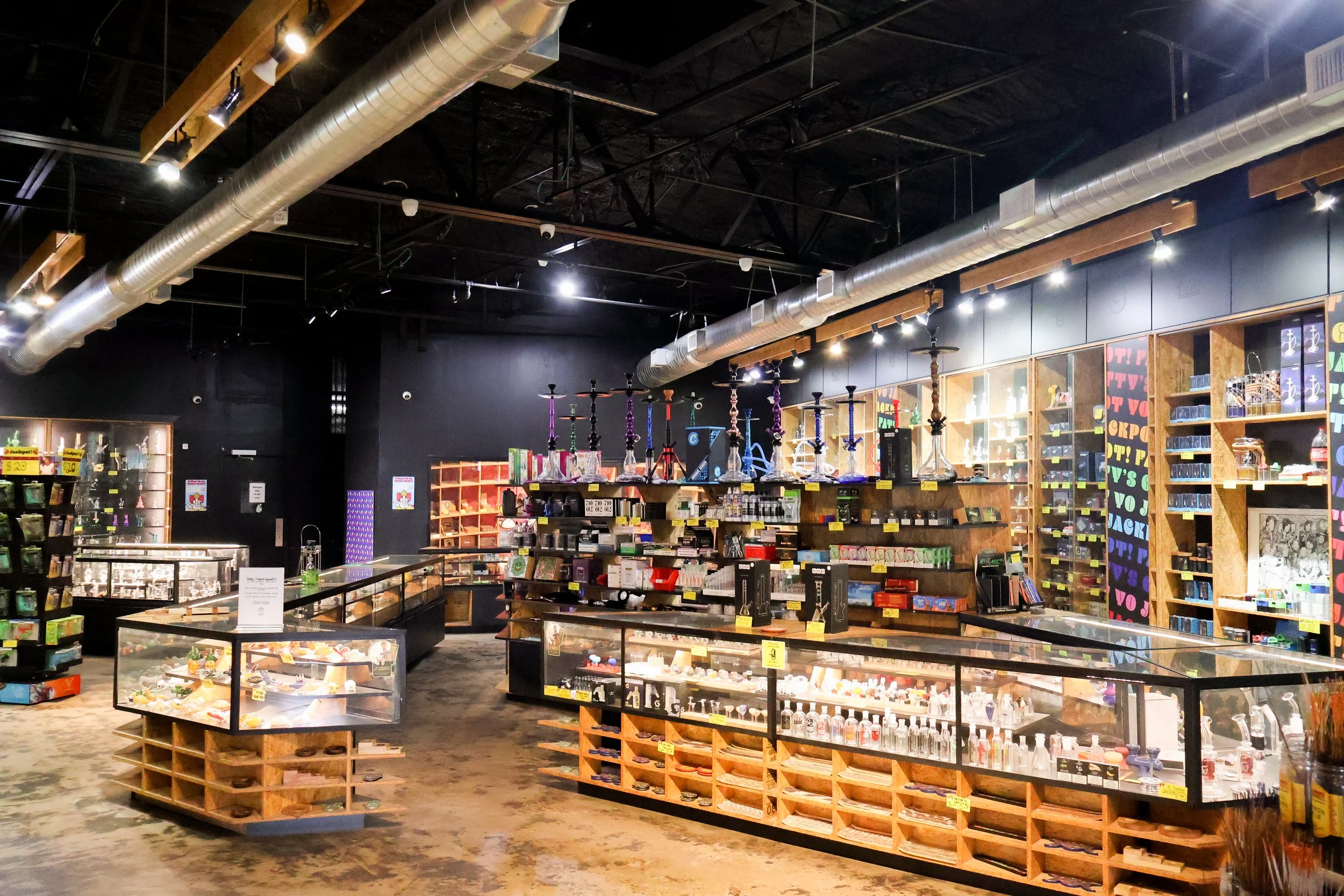 Shop interior with wooden display shelves holding various smoking accessories and legal paraphernalia. Hookah pipes are displayed on top of a central shelf, with shelves in the background stocked with other items. The ceiling features exposed ductwork and lighting.