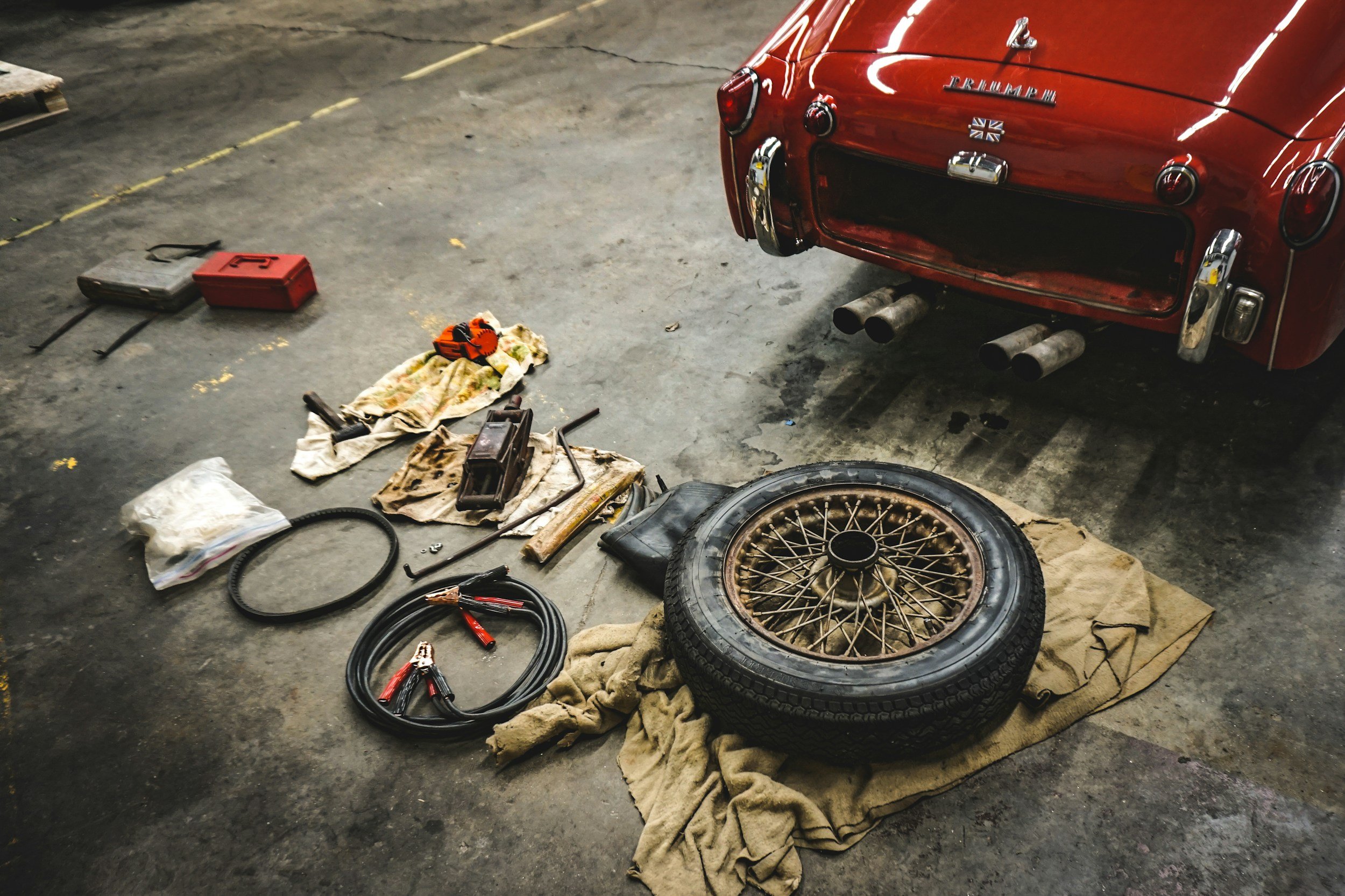 Inside a garage, a red Triumph classic car's rear end is elevated, with tools, a tire, and car parts scattered on the floor around it.