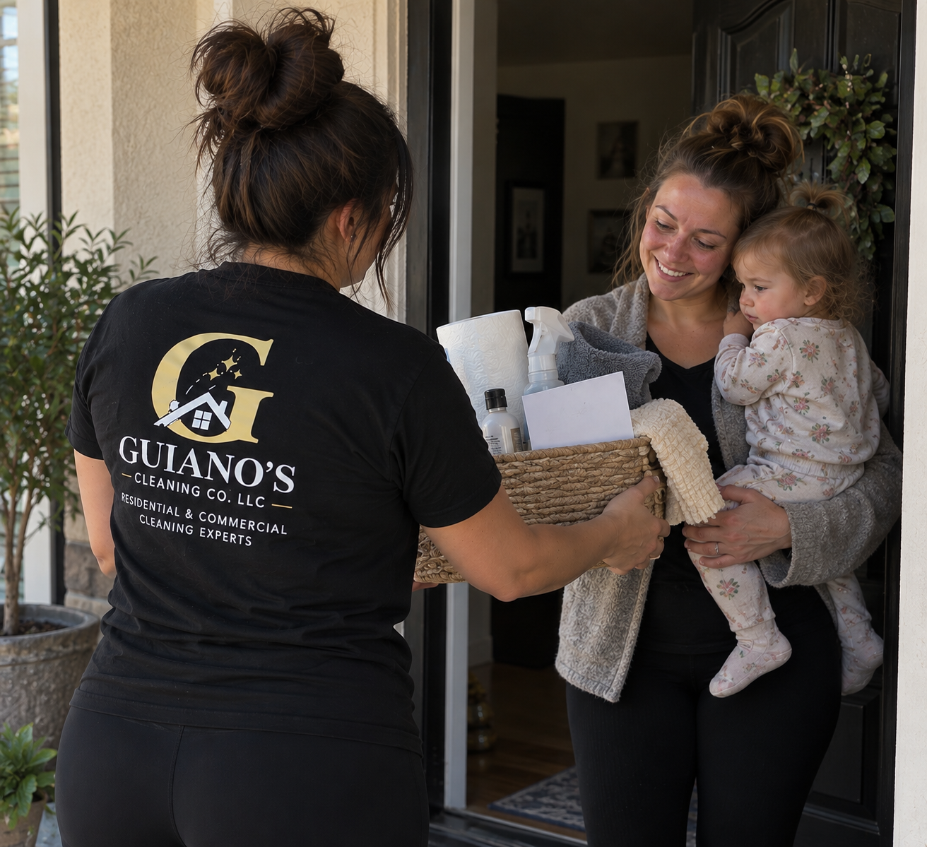A woman receives a basket of cleaning supplies as a gift from a cleaning service worker while standing at her front door, holding a young girl in pajamas. The cleaning service worker is seen from the back, wearing a black shirt with "Guano's Cleaning Co. LLC" logo.