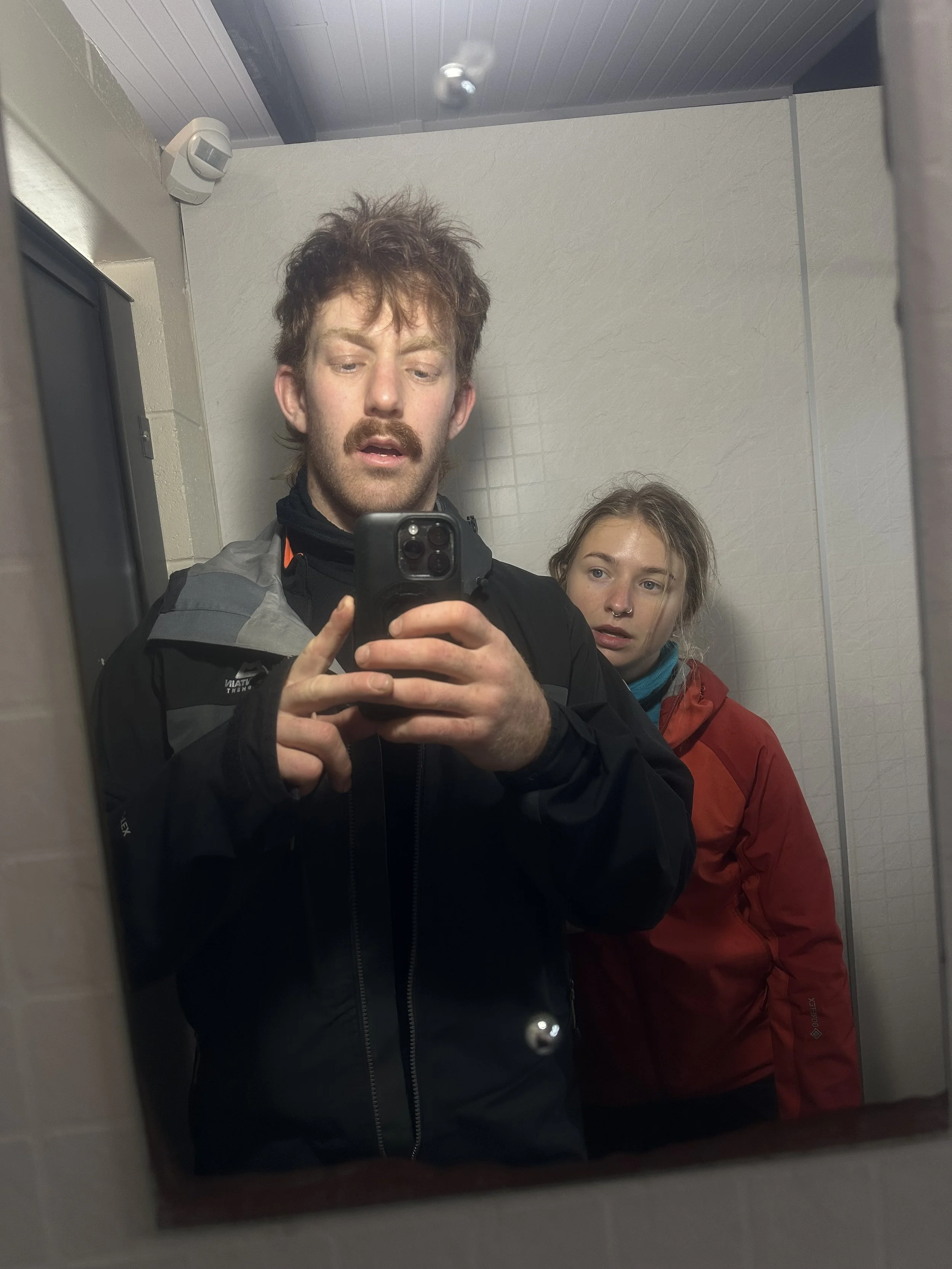 Young Aadventure couple stood taking selfie in mirror looking very tired and worn out in their waterproofs from their journeys.