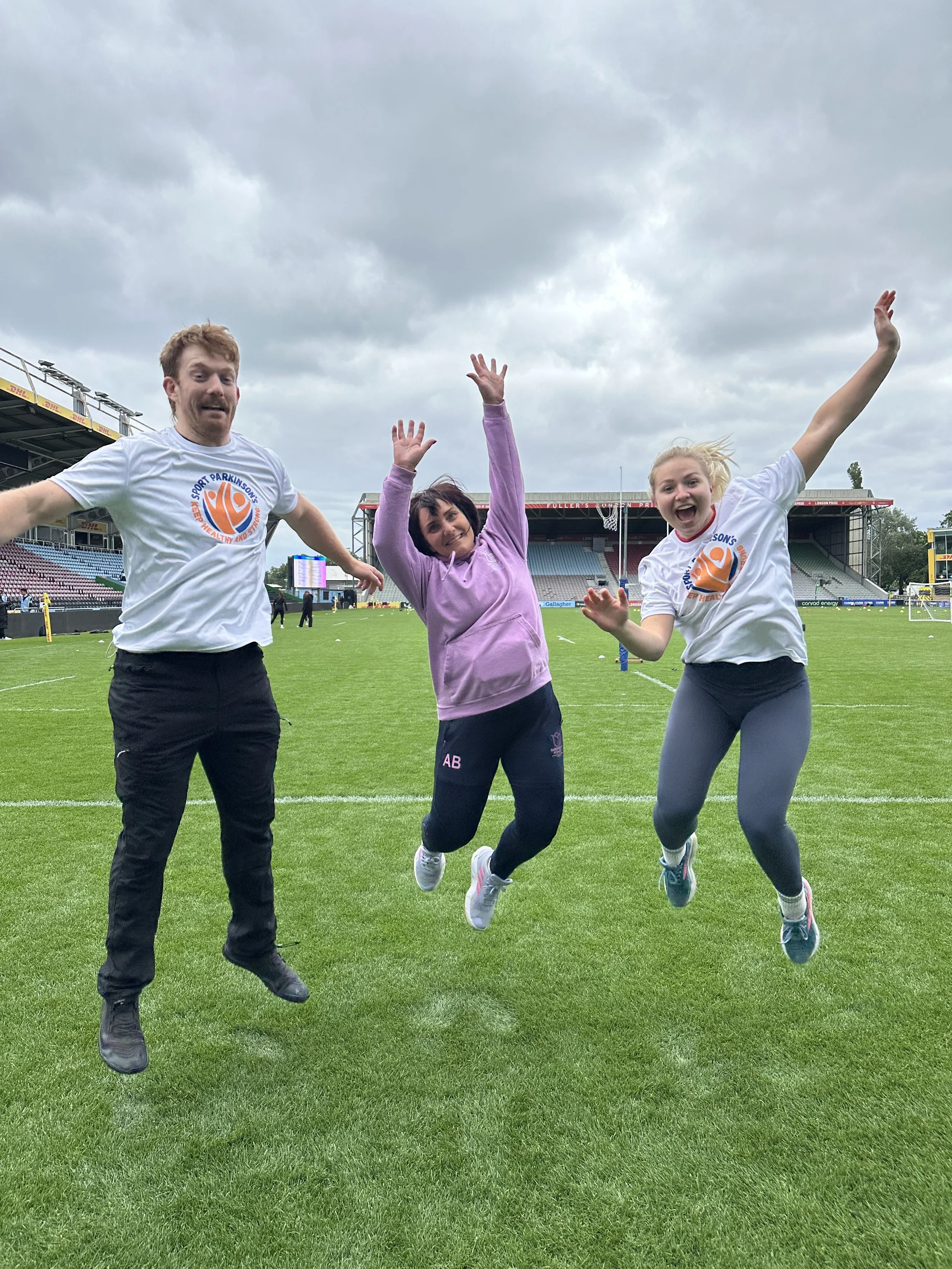 Three people in Sport Parkinson's T shirts jumping in the air at Twickenham stoop stadium. Charity event for Parkinson's disease. Two women and one man are smiling and waving.