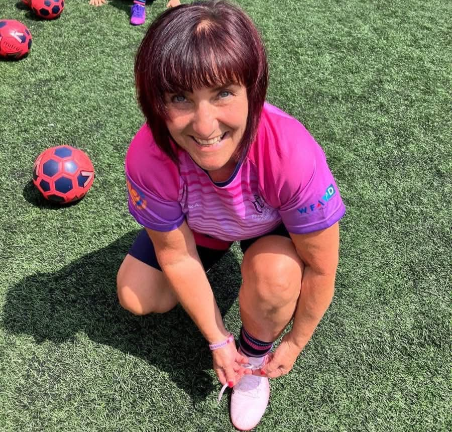 A woman in a pink sports jersey kneeling on a synthetic grass sports field, tying her shoelace, with soccer balls nearby, smiling at the camera.