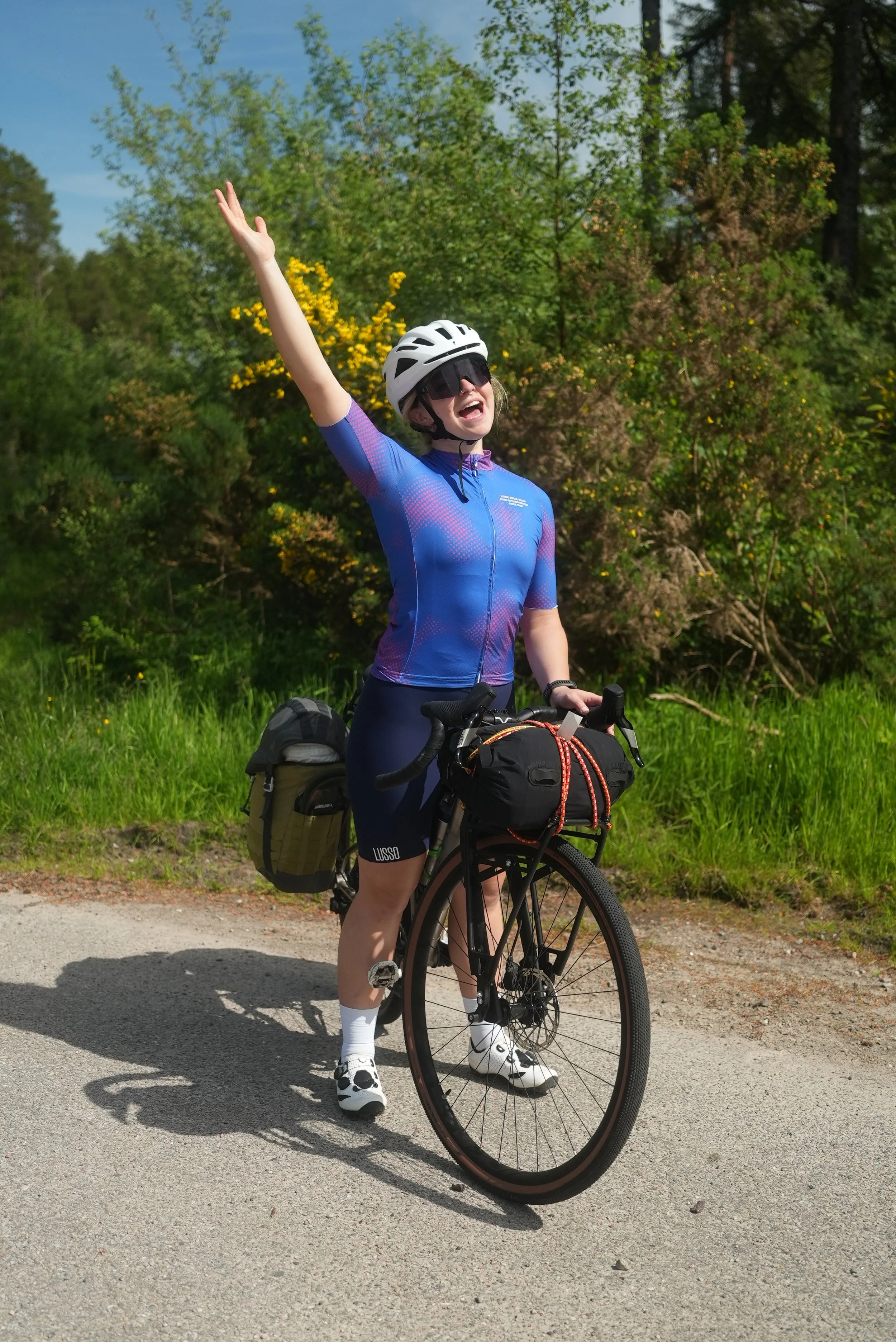 A woman wearing a cycling helmet, sunglasses, and a blue sports outfit standing next to her touring bicycle on a paved path, with green trees and bushes in the background, smiling and arms raised in a joyful pose.
