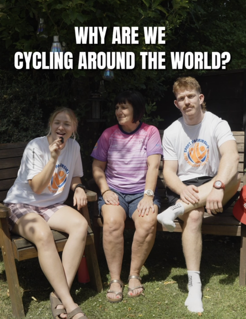 Three people sitting on a wooden bench outdoors, with a background of green foliage and hanging lights. The caption reads 'Why are we cycling around the world?'