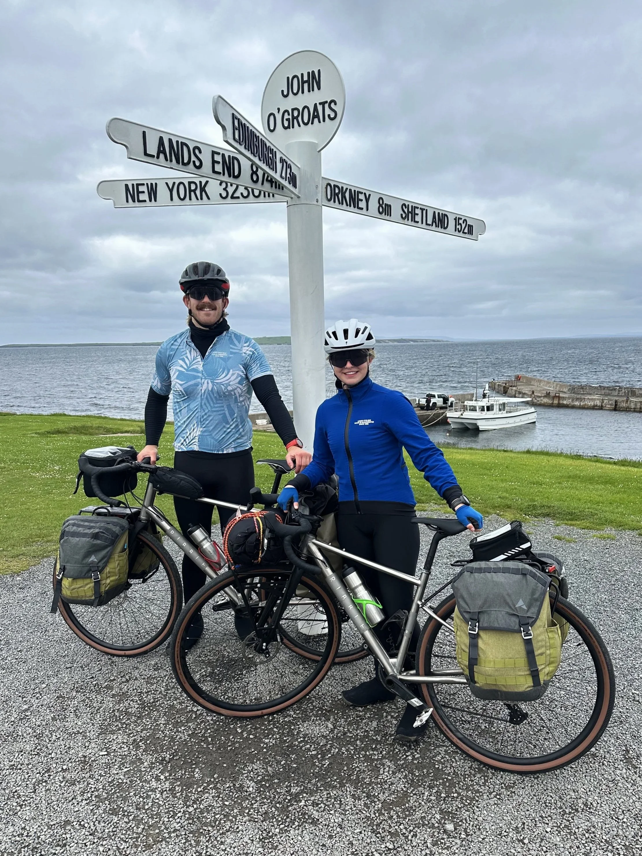 Two bikepackers stood with loaded panniers next to the John O'Groats sign by the sea in the Scottish Highlands
