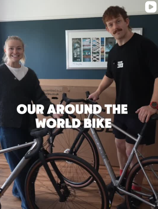 A woman and a man standing indoors with a black bicycle. The woman has blonde hair tied back and is smiling, wearing a dark sweater and jeans. The man has curly hair and a mustache, wearing a black t-shirt and dark shorts, holding the bicycle. There is a framed picture on the teal wall behind them and a cardboard box on the floor. Text on the image reads "OUR AROUND THE WORLD BIKE."