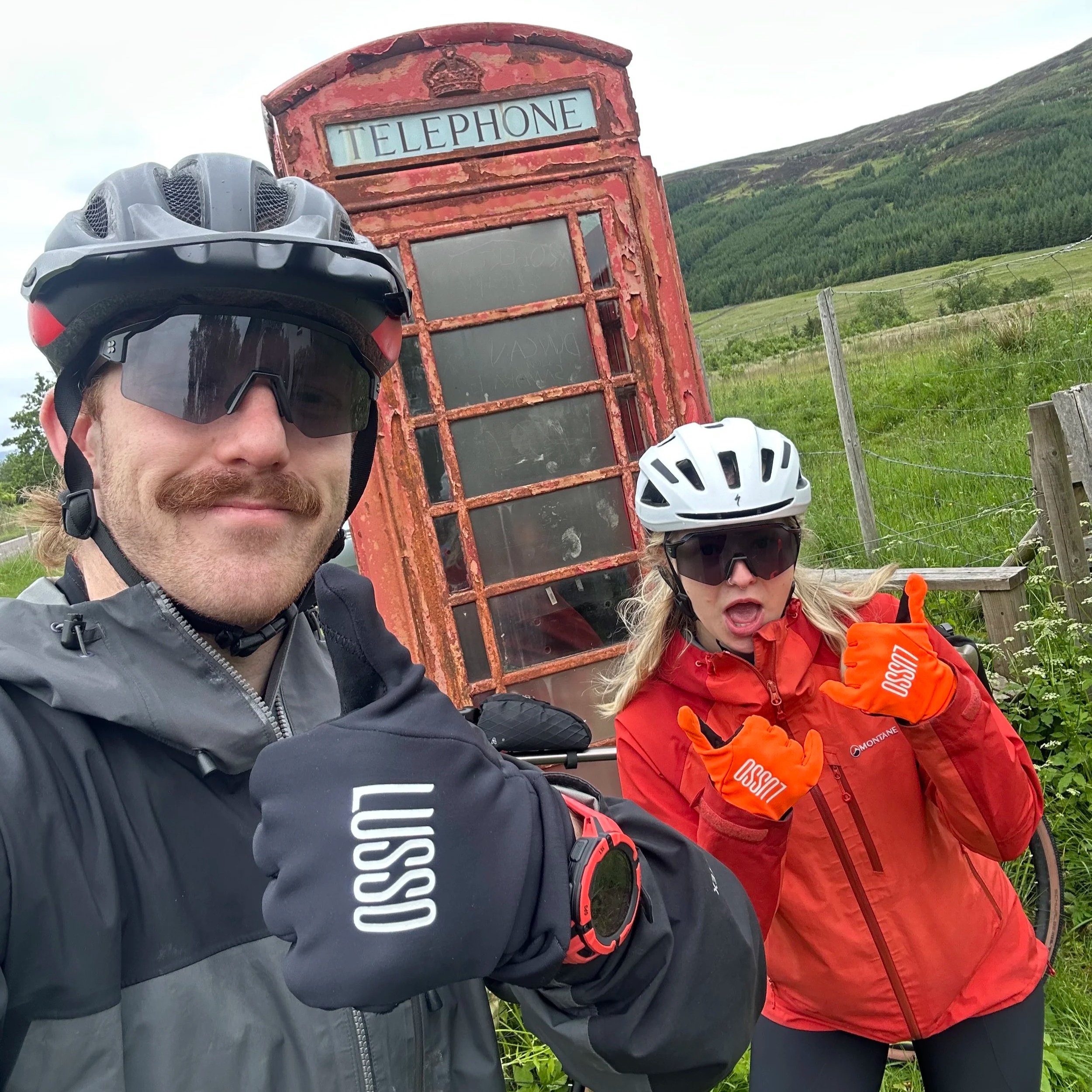 Two cyclists in helmets and jackets standing outdoors near a rusty red telephone booth with greenery and hills in the background.