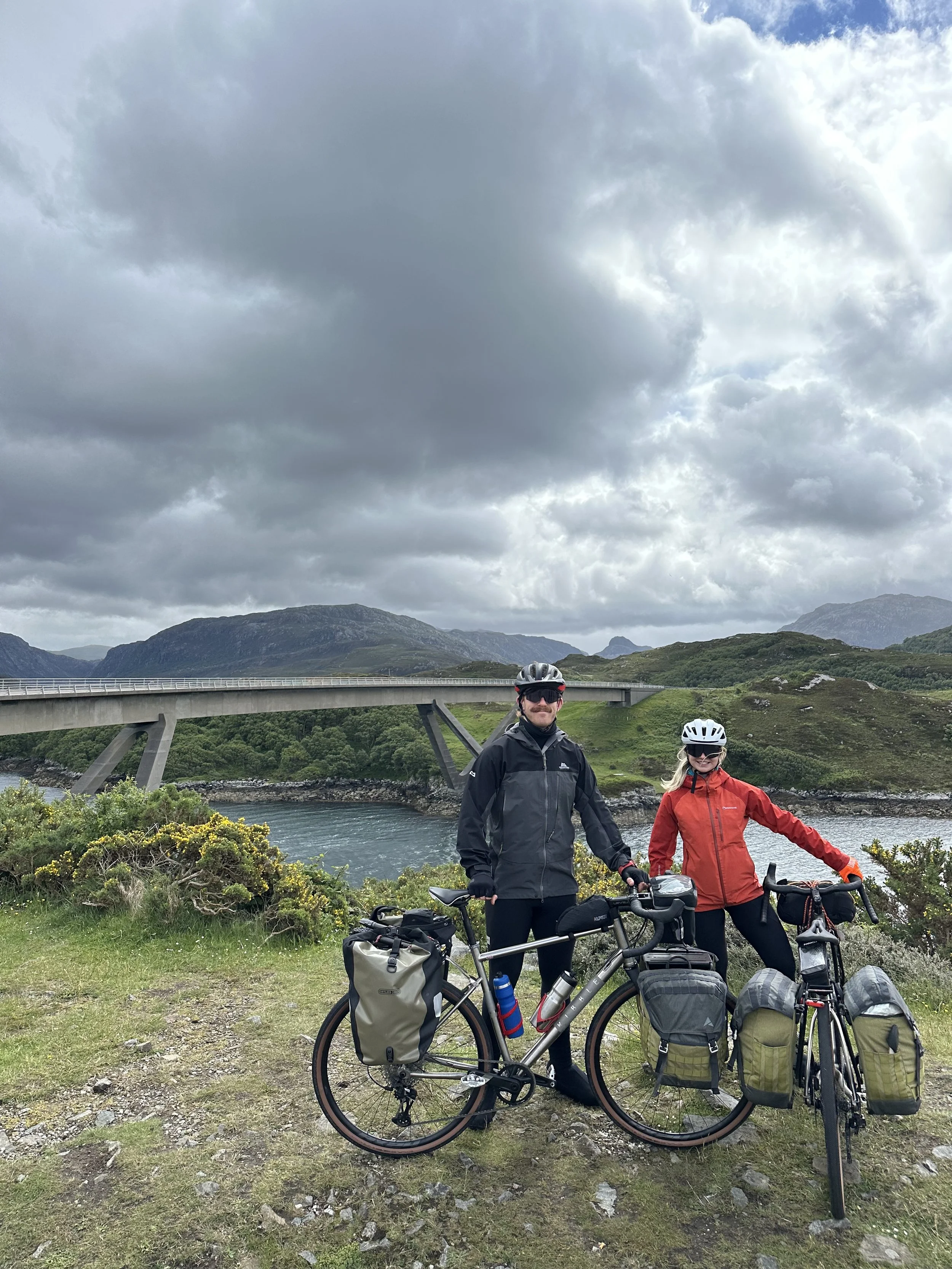 Two cyclists in outdoor gear standing next to their loaded bicycles near a river , with the Scottish mountains and the Kylesku bridge in the background under cloudy skies.