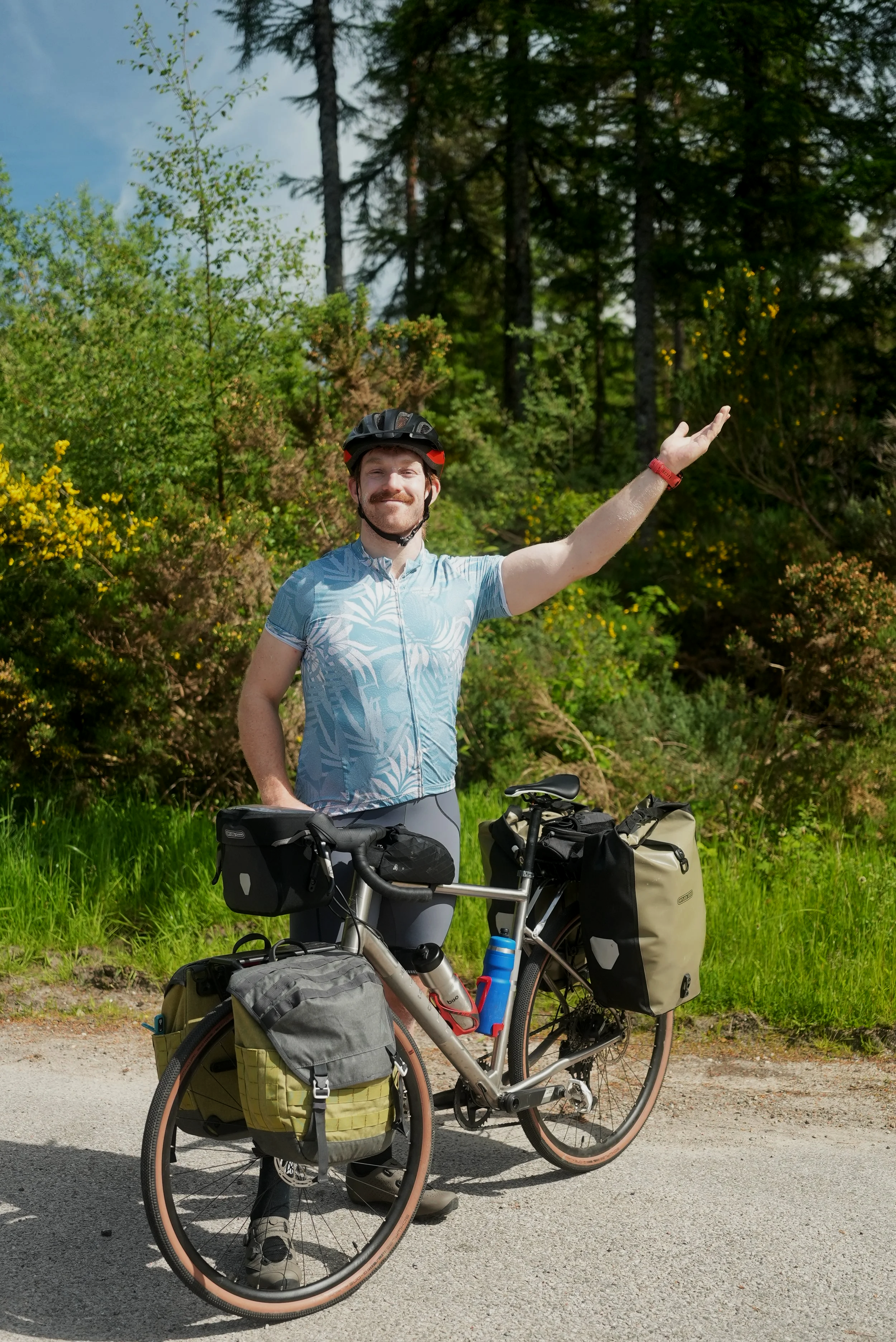 Smiling man in cycling gear and helmet standing next to fully loaded touring bicycle on a paved path in a forested area.
