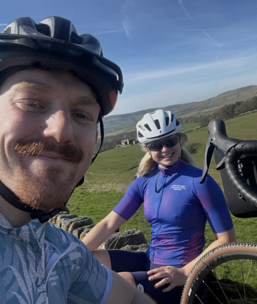 A man and woman in cycling gear and helmets smiling outdoors in a green field with hills in the background.